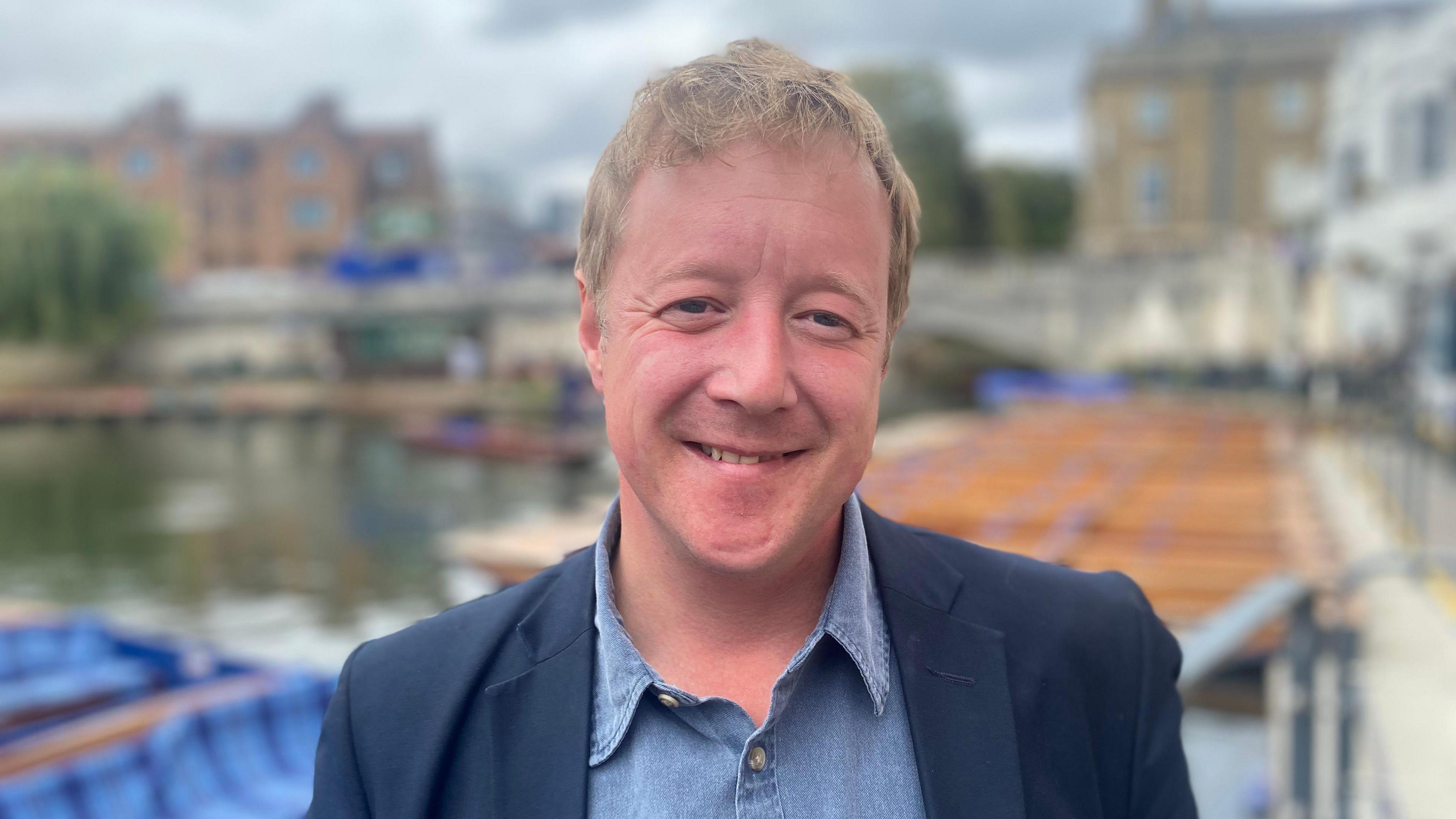 Paul Bristow, centre frame, smiling and looking directly down the camera. He is wearing a dark blue jacket and lighter blue, opened necked shirt. He is standing outside. The background is blurred, but a waterfront or riverside area and boats can be seen behind Mr Bristow.  