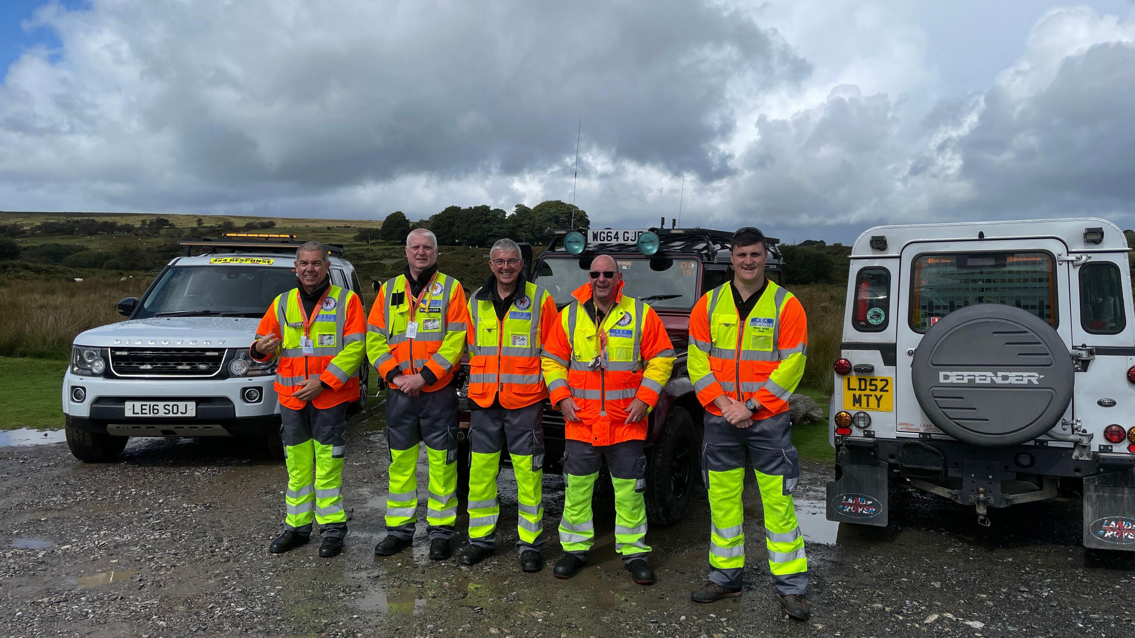 Five men stood shoulder-to-shoulder in front of three 4x4 vehicles. They are all wearing bright yellow and orange, high-viz clothing.