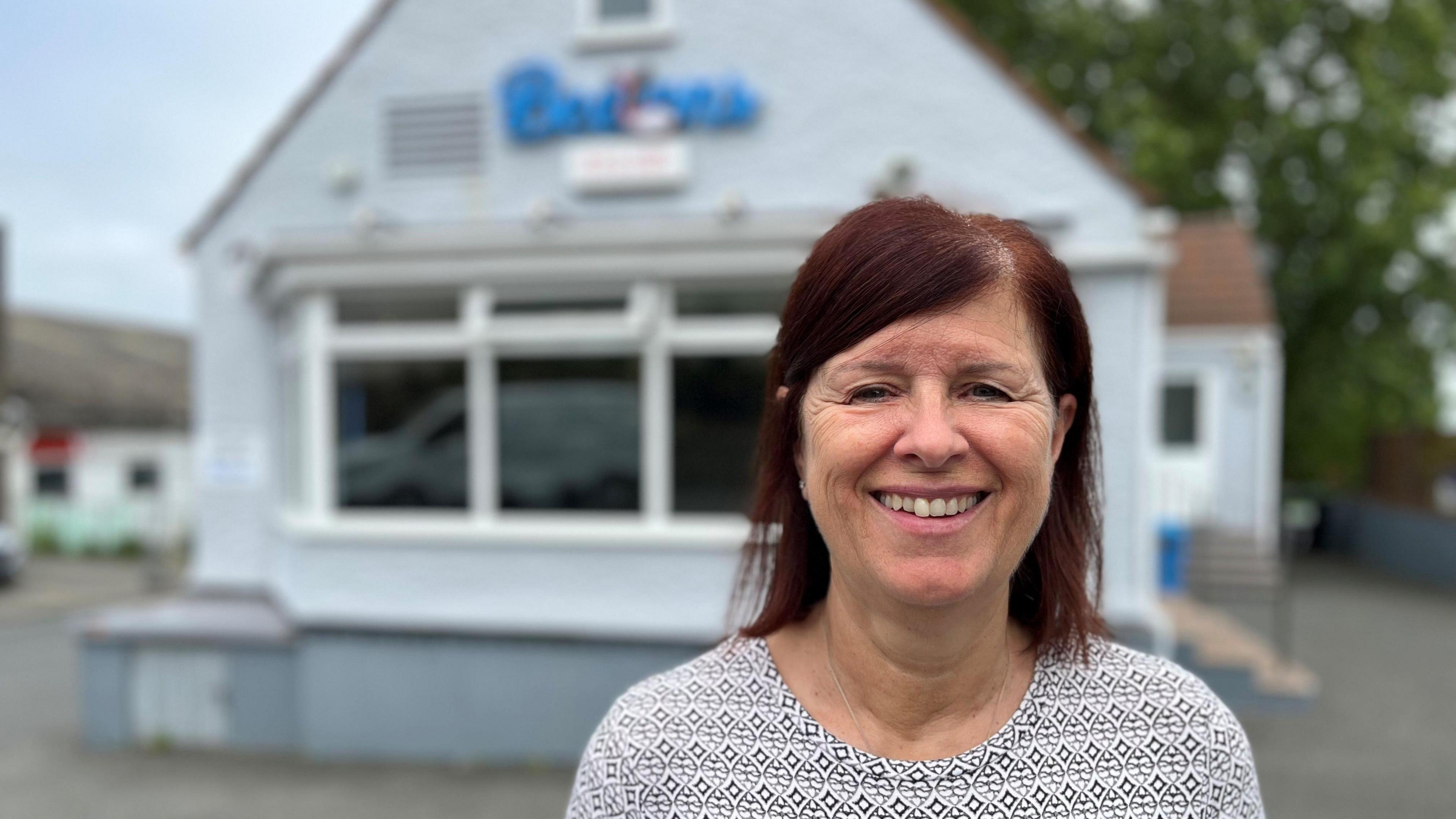 Kim Ashplant smiling at looking at the camera. She is wearing a light coloured top and has dark red hair. She is stood in front of the fish and chip shop which is blurred.