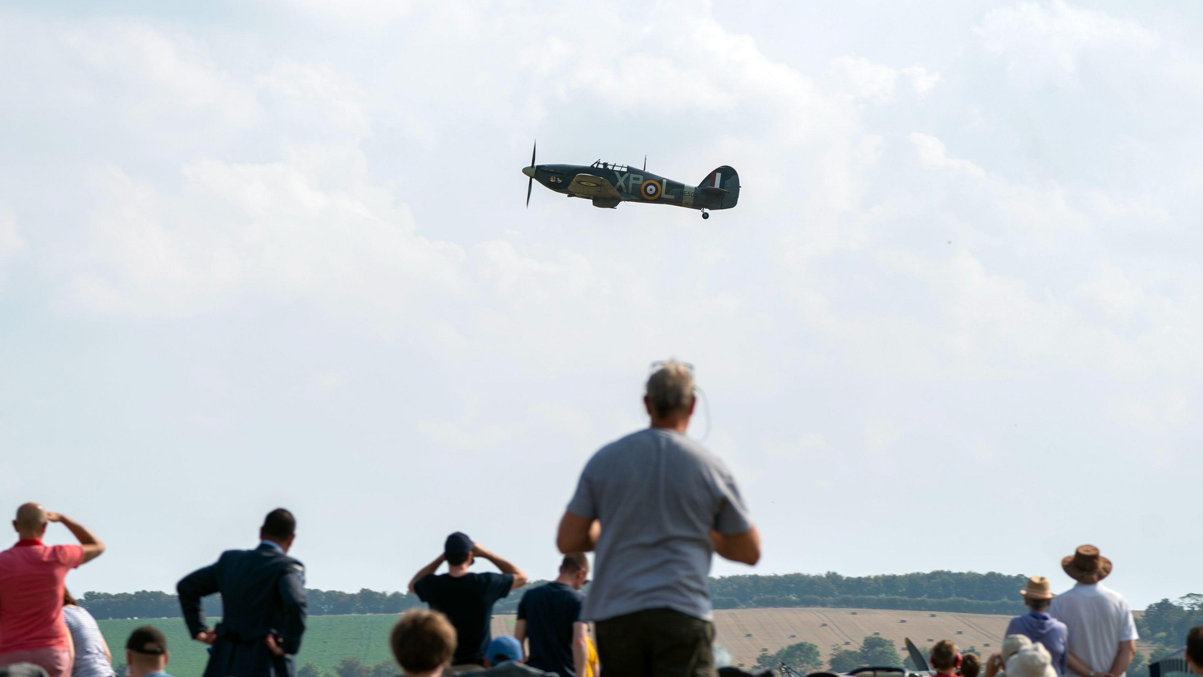 A photo of crowds watching a Hawker Hurricane at a different event on a sunny and clear day. The aircraft is predominately dark green with a yellow, blue, white and red randall on the tail.