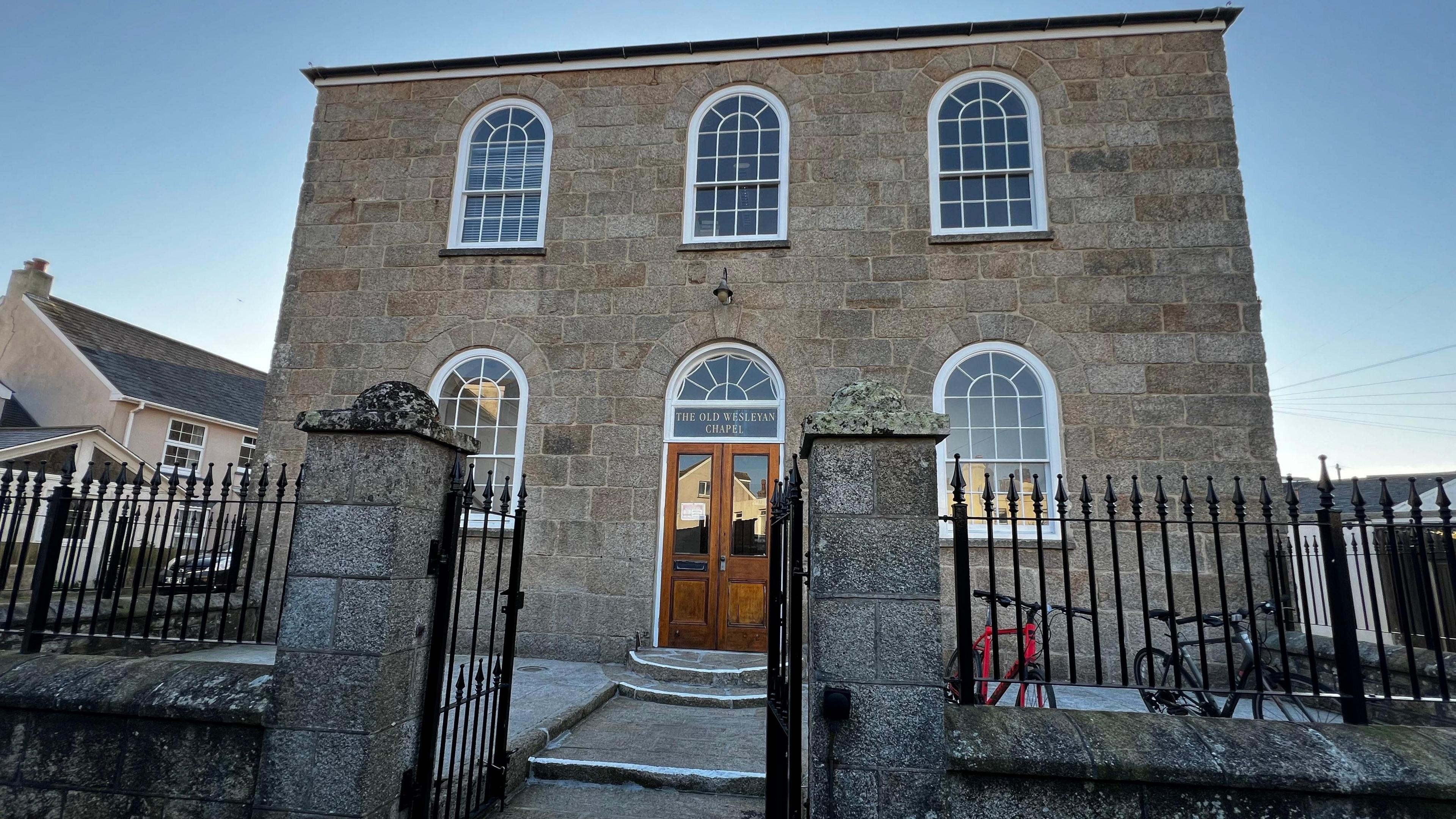 A picture of the stone building at dusk. There are arched windows with a set of wooden double doors and a path leading up to the entrance. A black metal wrought iron fence surrounds the perimeter.