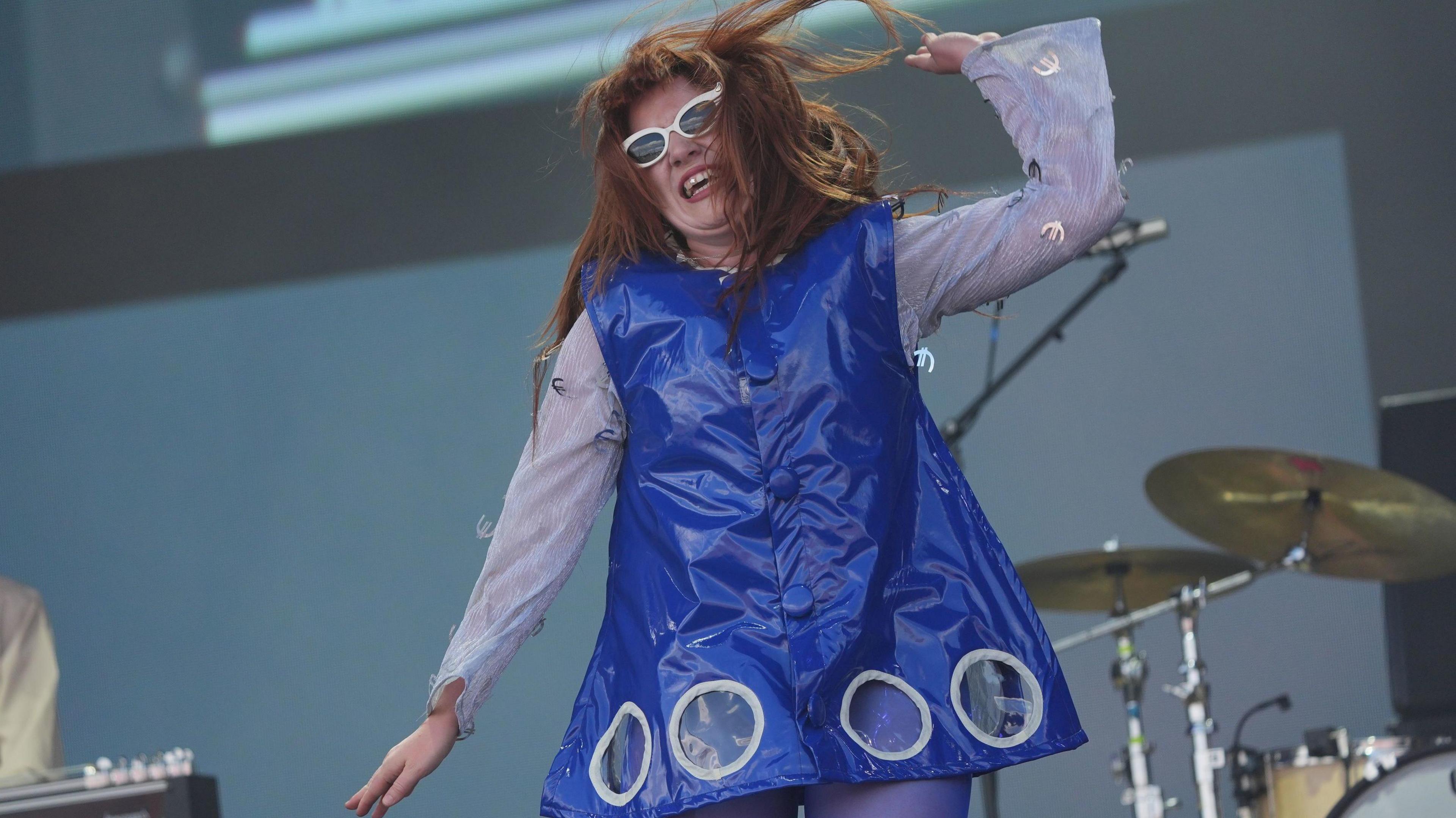 Musician CMAT in a blue dress dancing on stage in front of a drum set.
