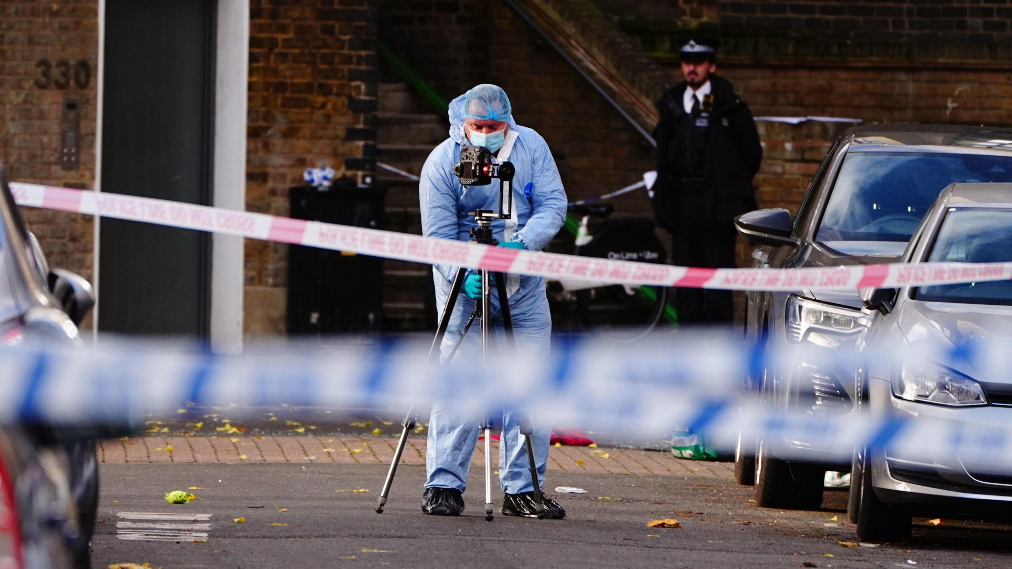 A police forensic officer in a blue protective suit, gloves and hairnet takes a photo of the crime scene behind a cordon on Southern Grove. A red cloth and green and yellow paramedic kit bag can be seen on the ground behind them