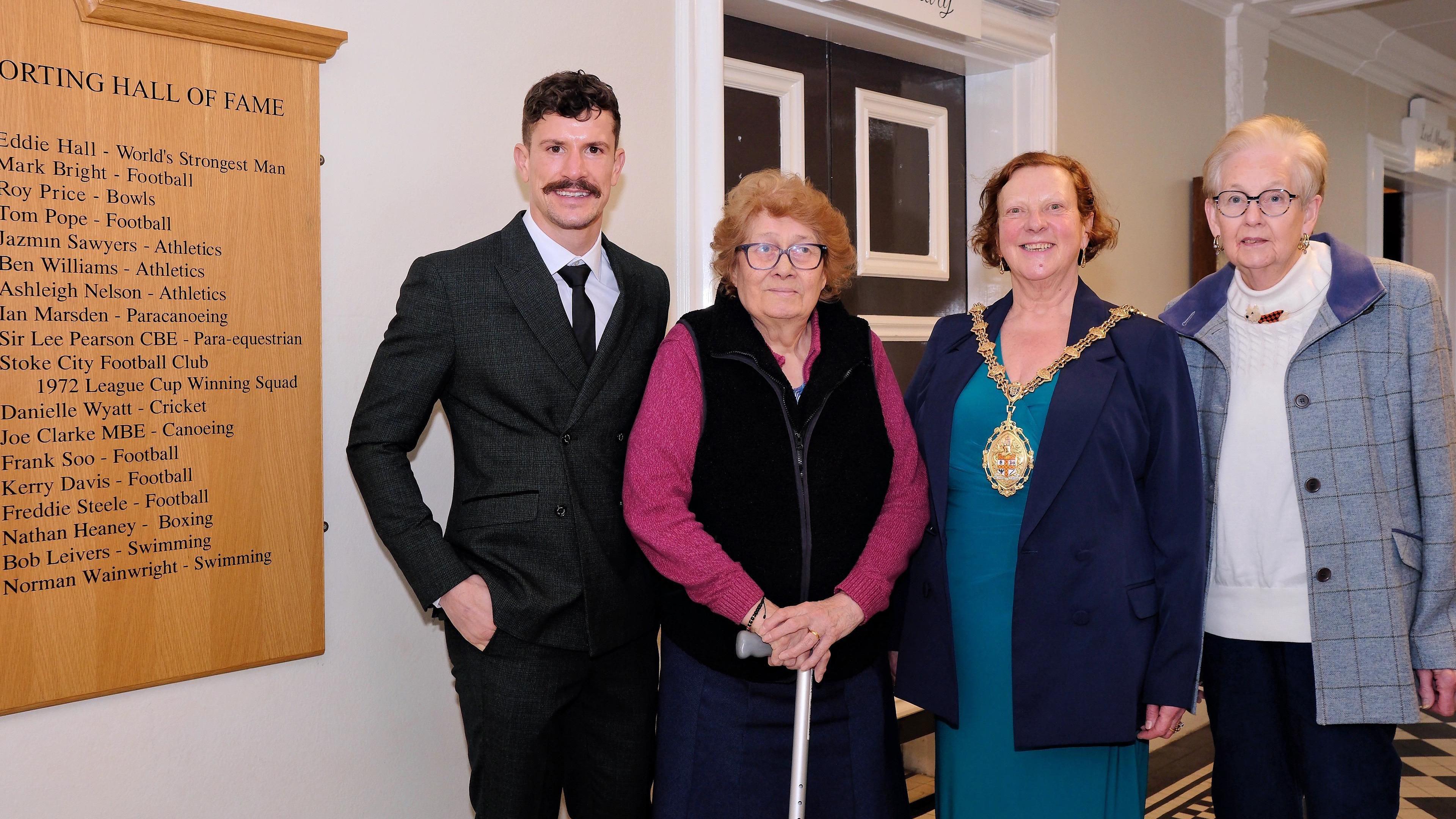 Nathan Heaney, wearing a black suit, is stood next to three women. He is pictured alongside a wooden board which lists sporting stars from Stoke-on-Trent.