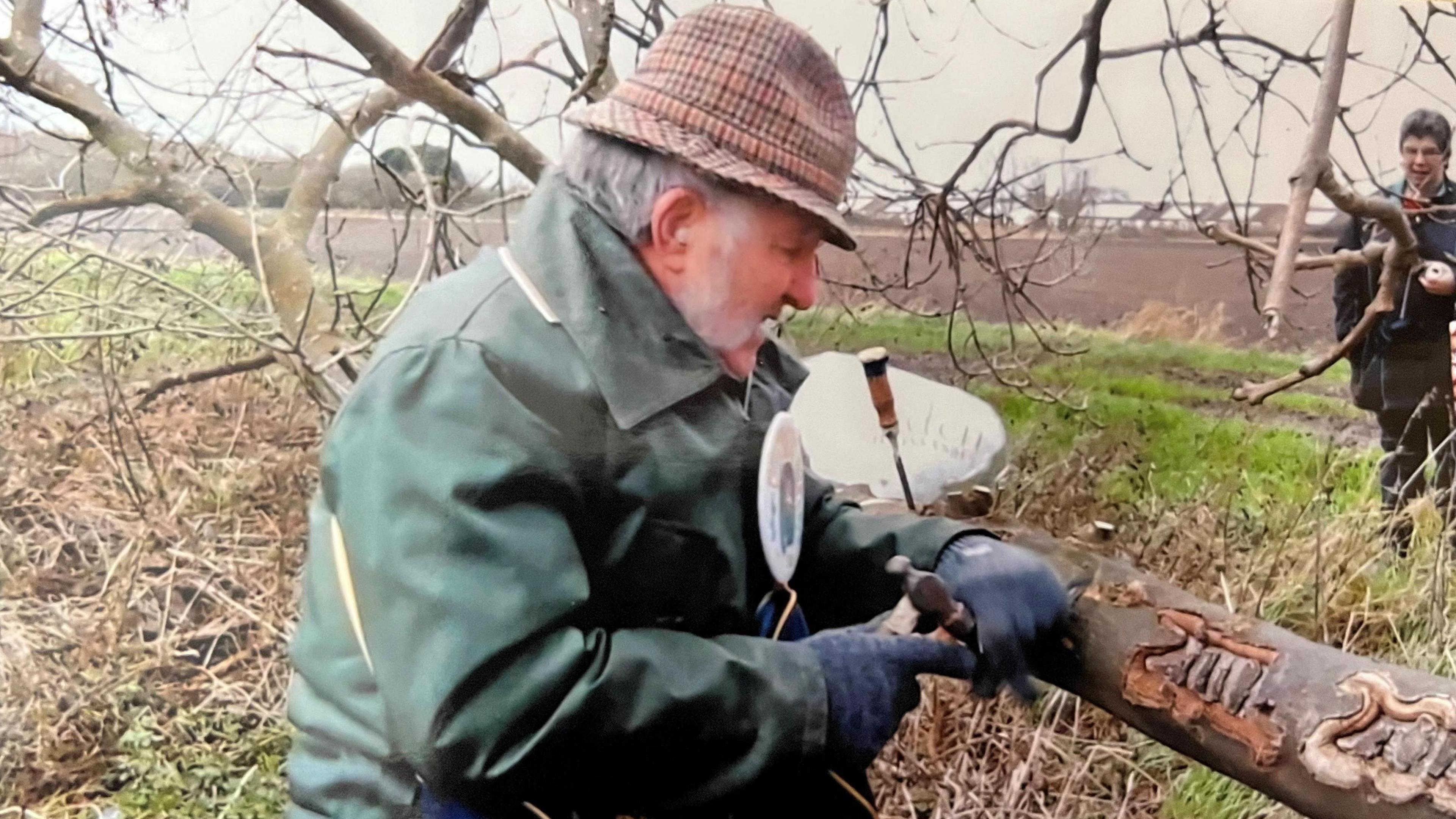 Colin Bedford, 88, wearing a green wax jacket overcoat, he has a tartan fedora hat and long grey whiskery sideburns, and has dark gloves, he is using a hammer and chisel to carve the year into the thick branch of an ash tree. Farmland is beyond him, a ploughed brown field with some trees in the distance, a person is watching him work to the right.