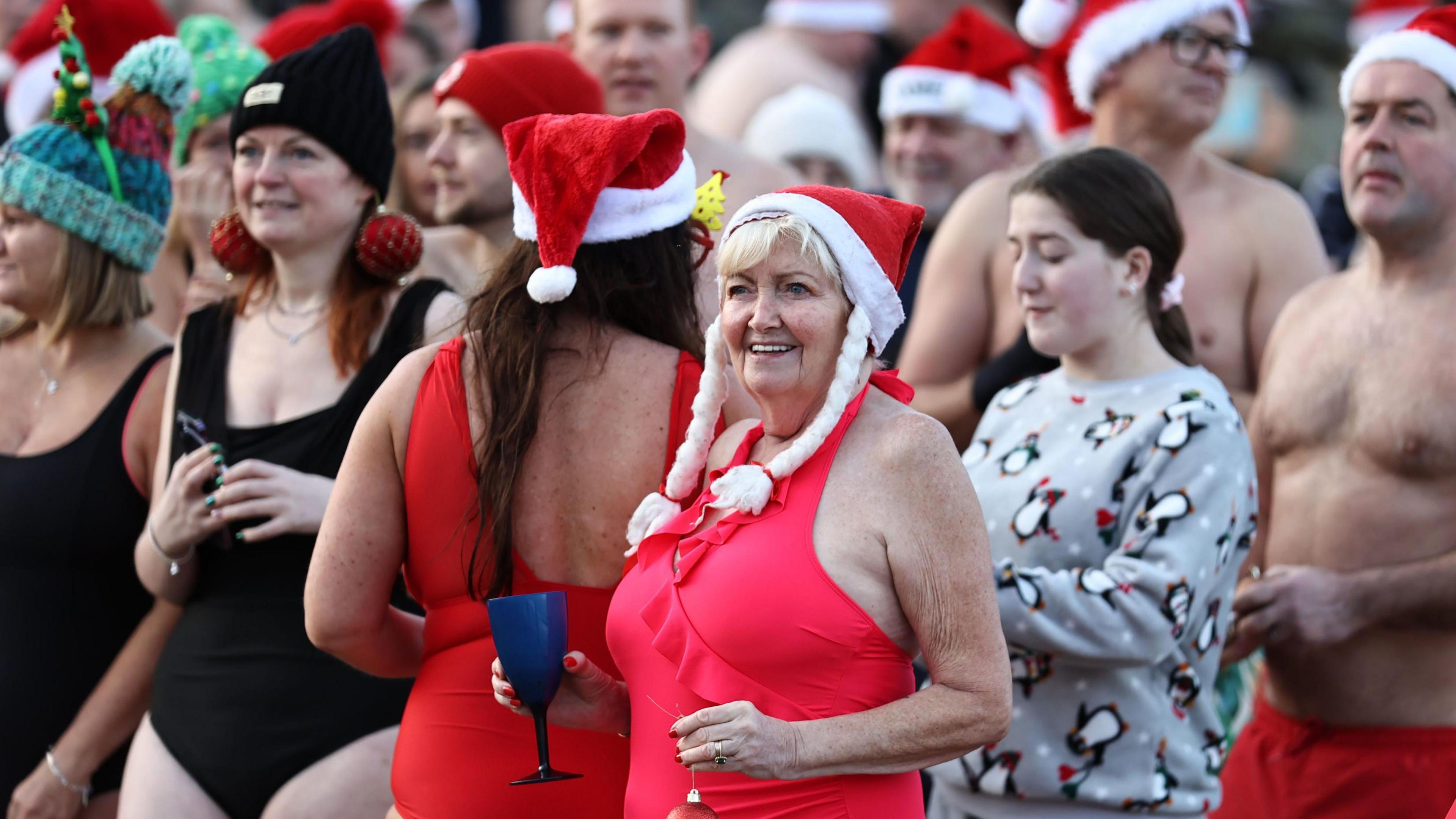 A woman in a red swimsuit holding a blue cup in one hand and a Christmas tree bauble in the other. She has a red Santa hat one which two long plaits are hanging out of.