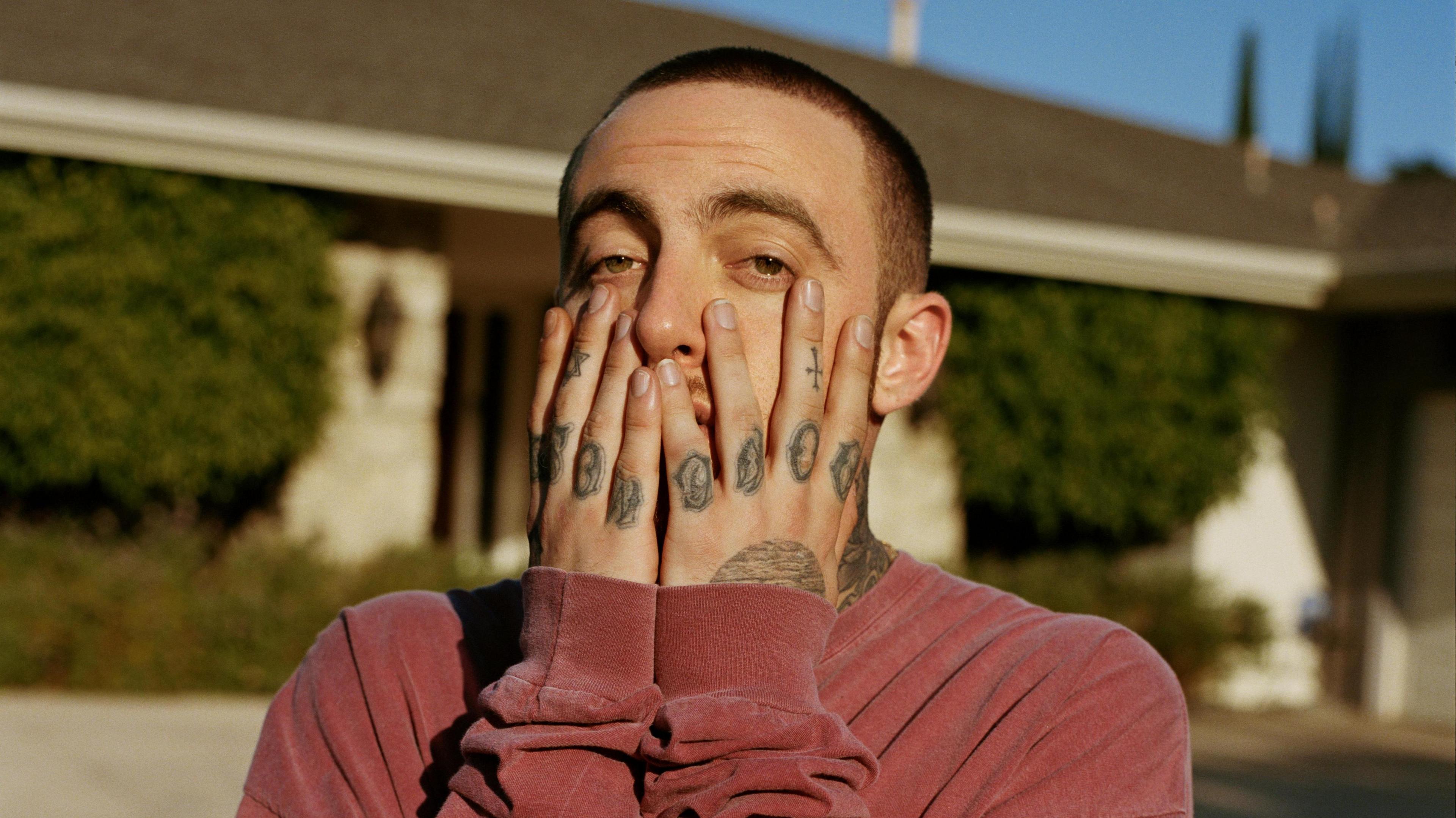 Mac Miller poses with his hands on his face for a magazine shoot. He's pictured outside a single-storey house on a blue-sky day. He has croppped brown hair, brown eyes and tattooed fingers and wears a red sweater. 