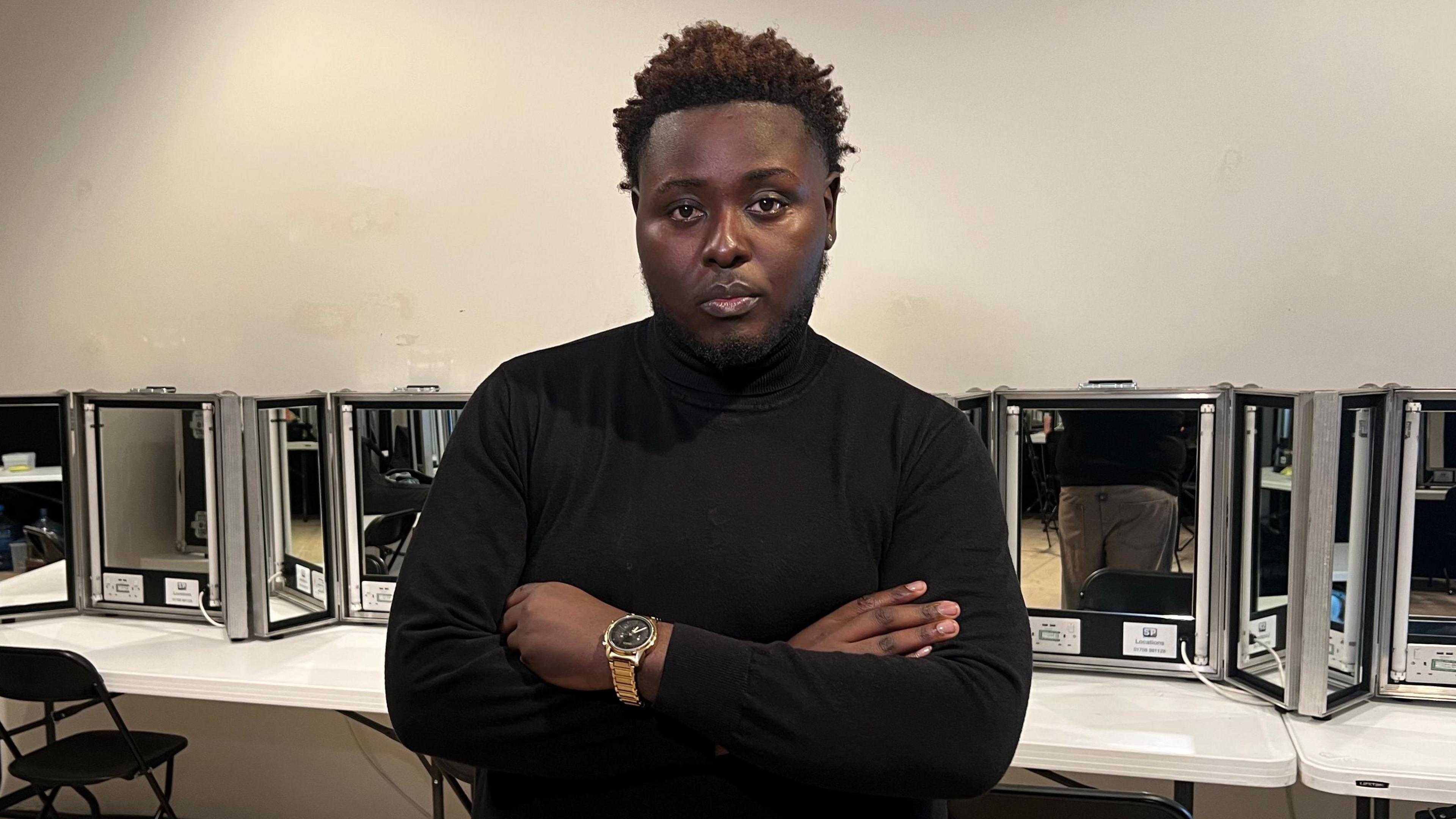 Model James Corbin stood in a dressing room. He is wearing a black jumper, with a gold watch on his left wrist. His arms are crossed and he is looking directly into the camera.