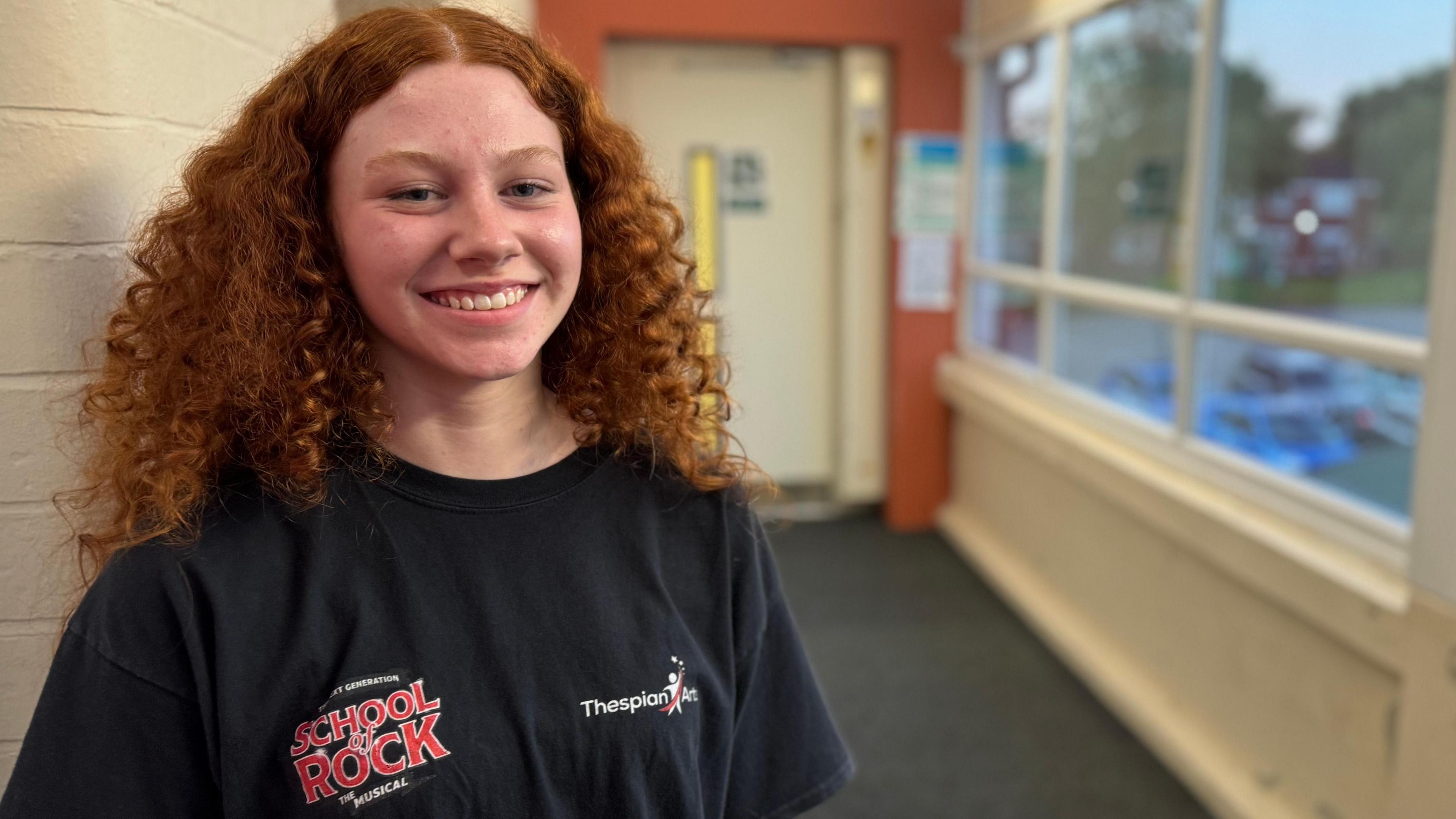 Annabel smiling at the camera. She has curly long hair and is wearing a black T-shirt with "School of Rock" written on it. 