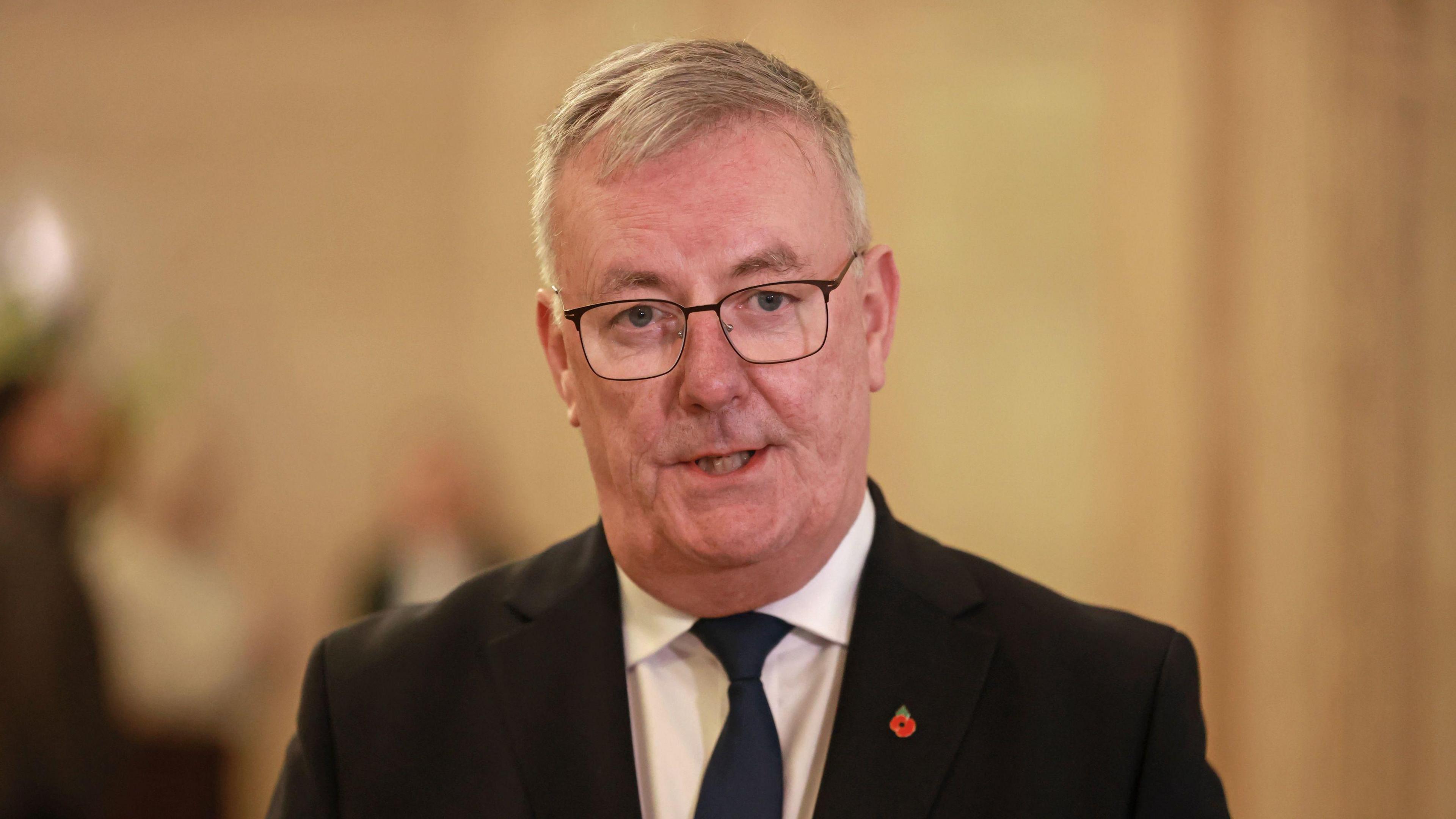 Mike Nesbitt wears a black suit and a white shirt with a navy tie. He has short white hair and wears glasses. 