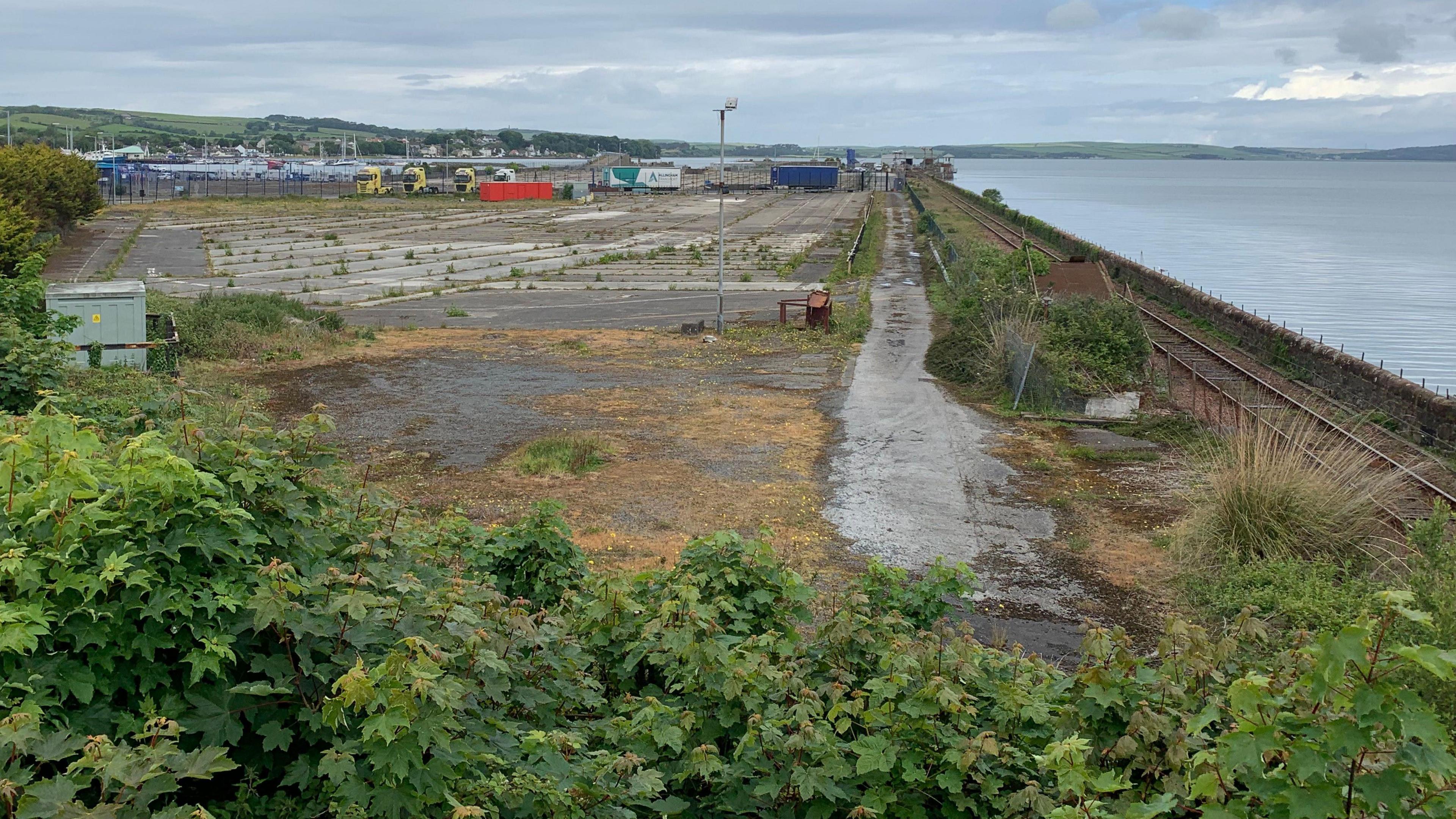 A slice of abandoned and overgrown land with a rail track down one side and the sea next to it with lorries in the distance