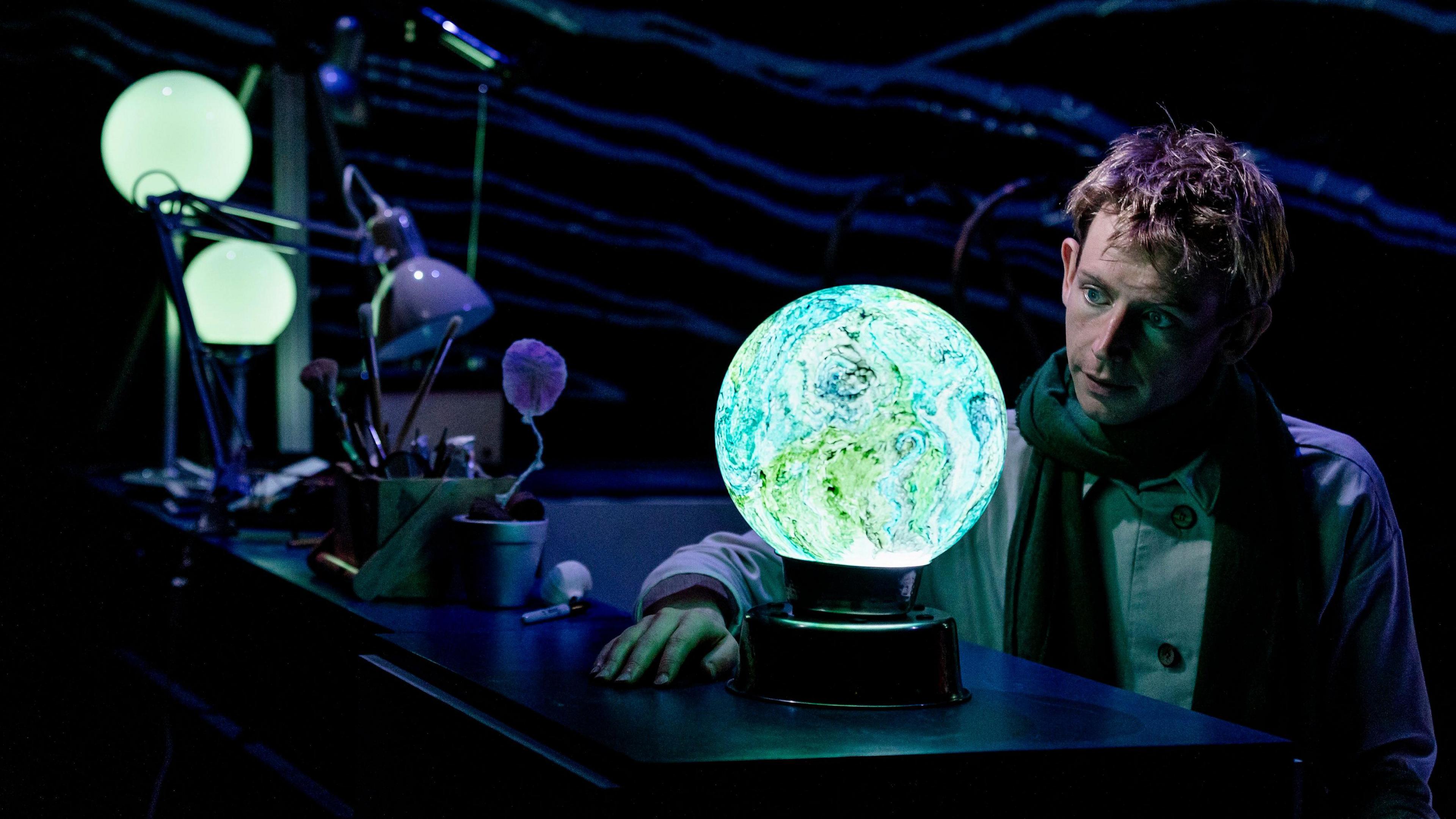 Toby Thompson performing The Little Prince at The Egg Theatre in Bath, Somerset. Toby has short dark blonde hair and is wearing a jacket with buttons and a scarf. The stage dark, but there are three lamps illuminating Toby's face. One of the lamps is a sphere and patterned blue and green, like the Earth. Toby is looking at it.