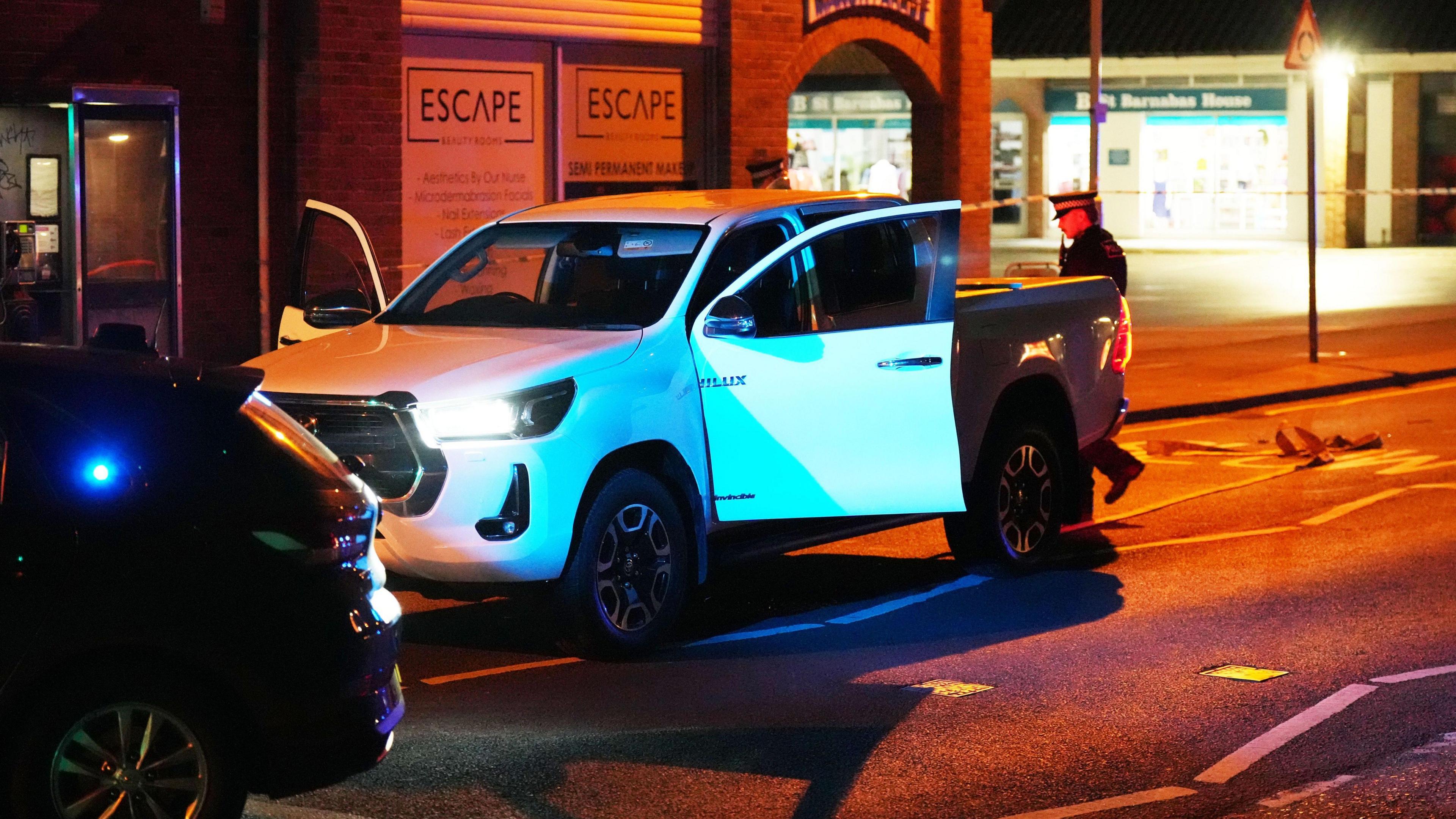 A white Toyota Hilux, a large pick up truck, used in a ram raid robbery, stands abandoned on the road in the darkness. Both its doors are open, as a police officer walks around the back of the vehicle.
