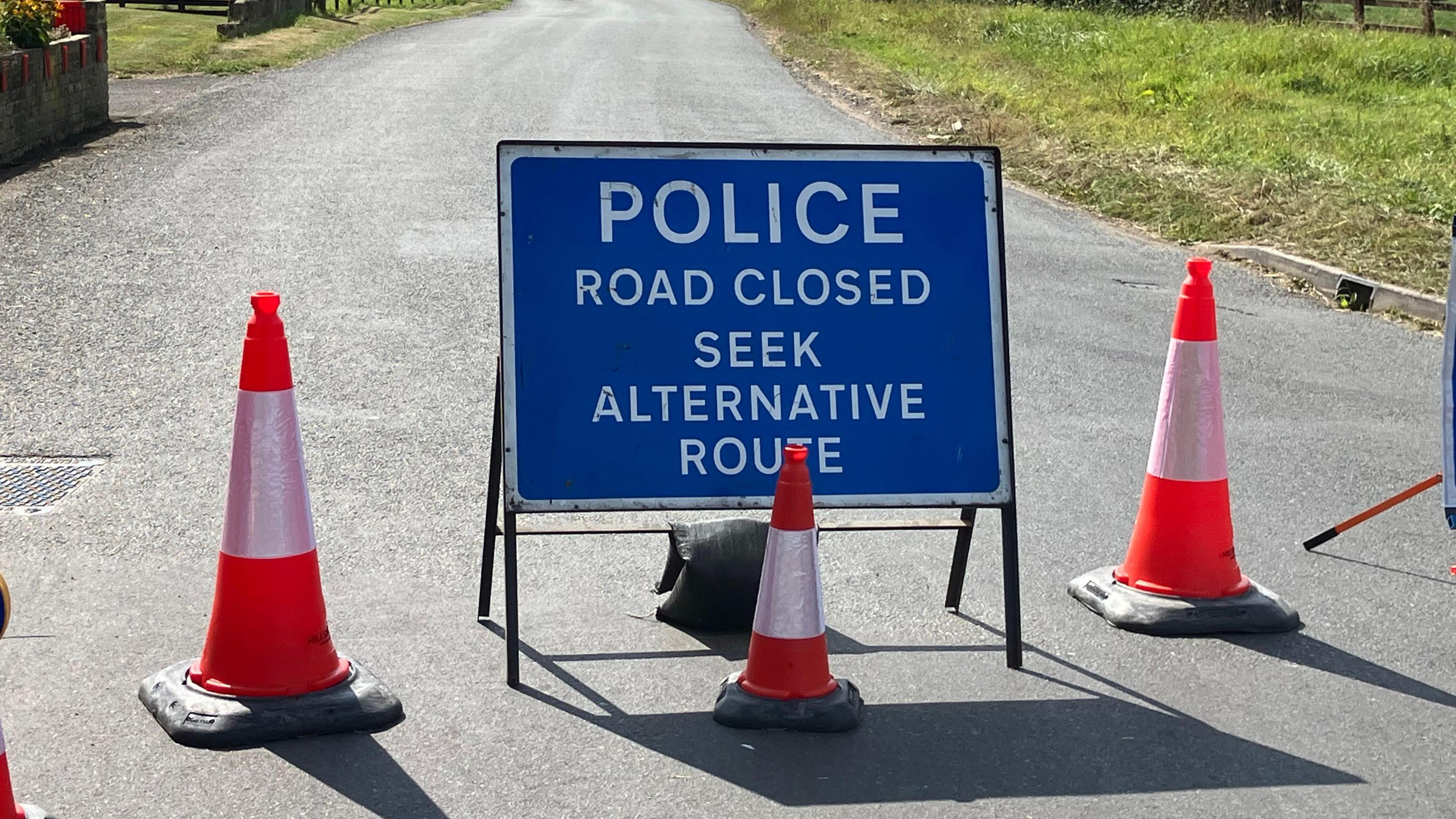 Blue police sign reads 'Police Road Closed Seek Alternative Route'. Sign is on country road and surrounded by traffic cones.