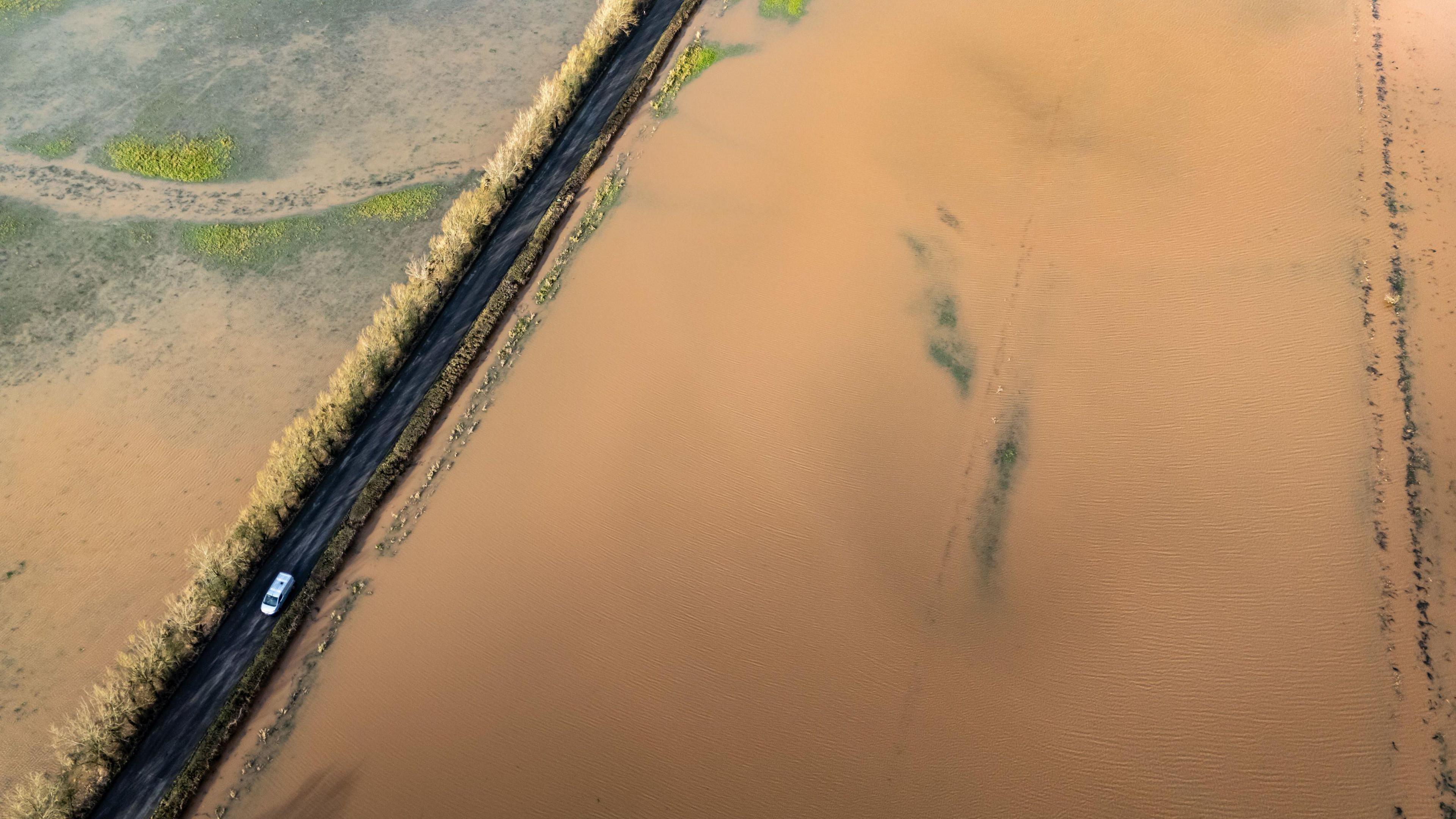 A birds-eye perspective of a flooded field. The water is brown. A van is driving on a road next to the field.