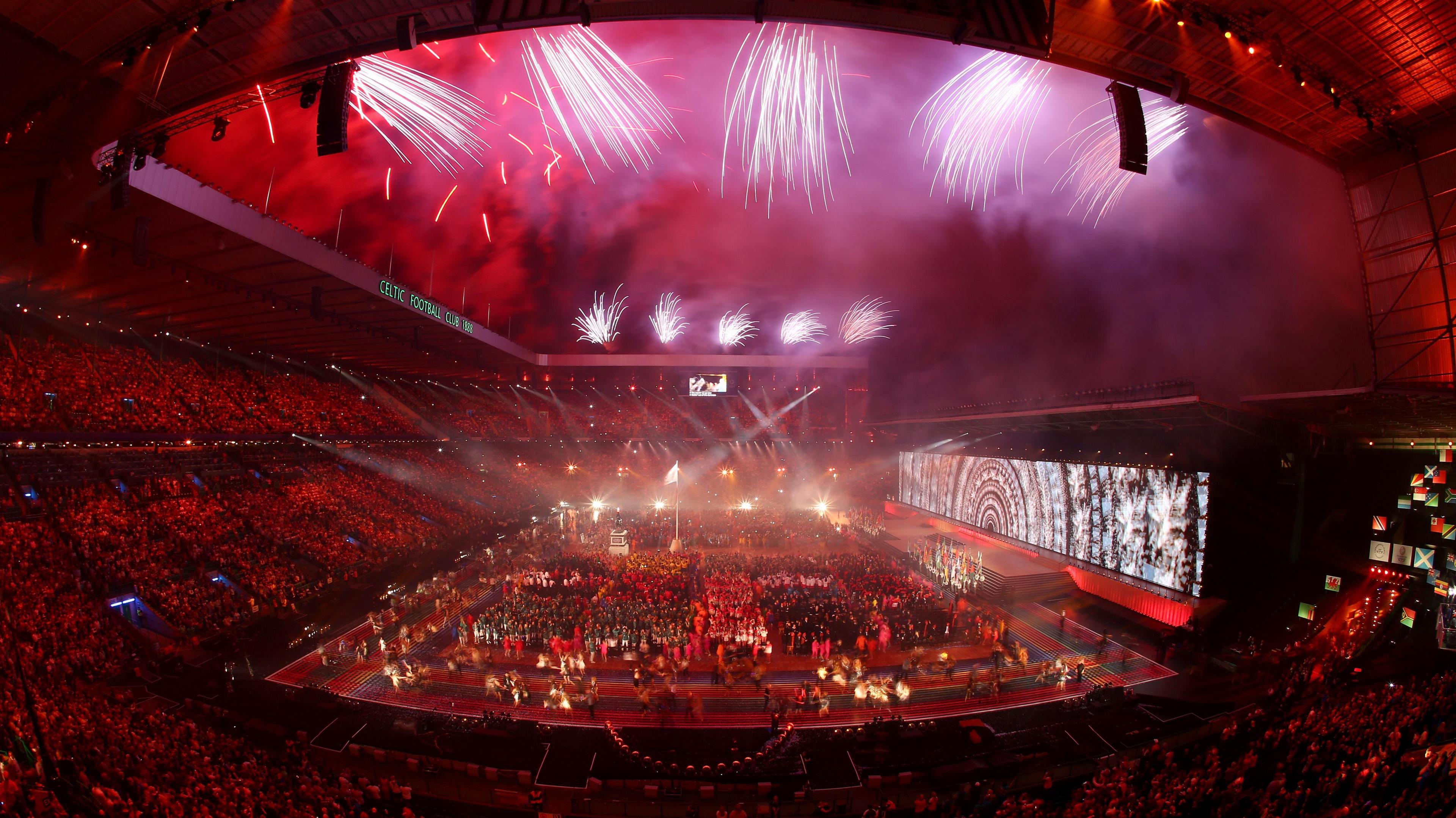 Fireworks illuminate the night sky over a stadium during the opening ceremony of the 2014 Glasgow Commonwealth Games. Thousands of people watch the action from the stands while hundreds of athletes stand on the pitch in front of a large screen.