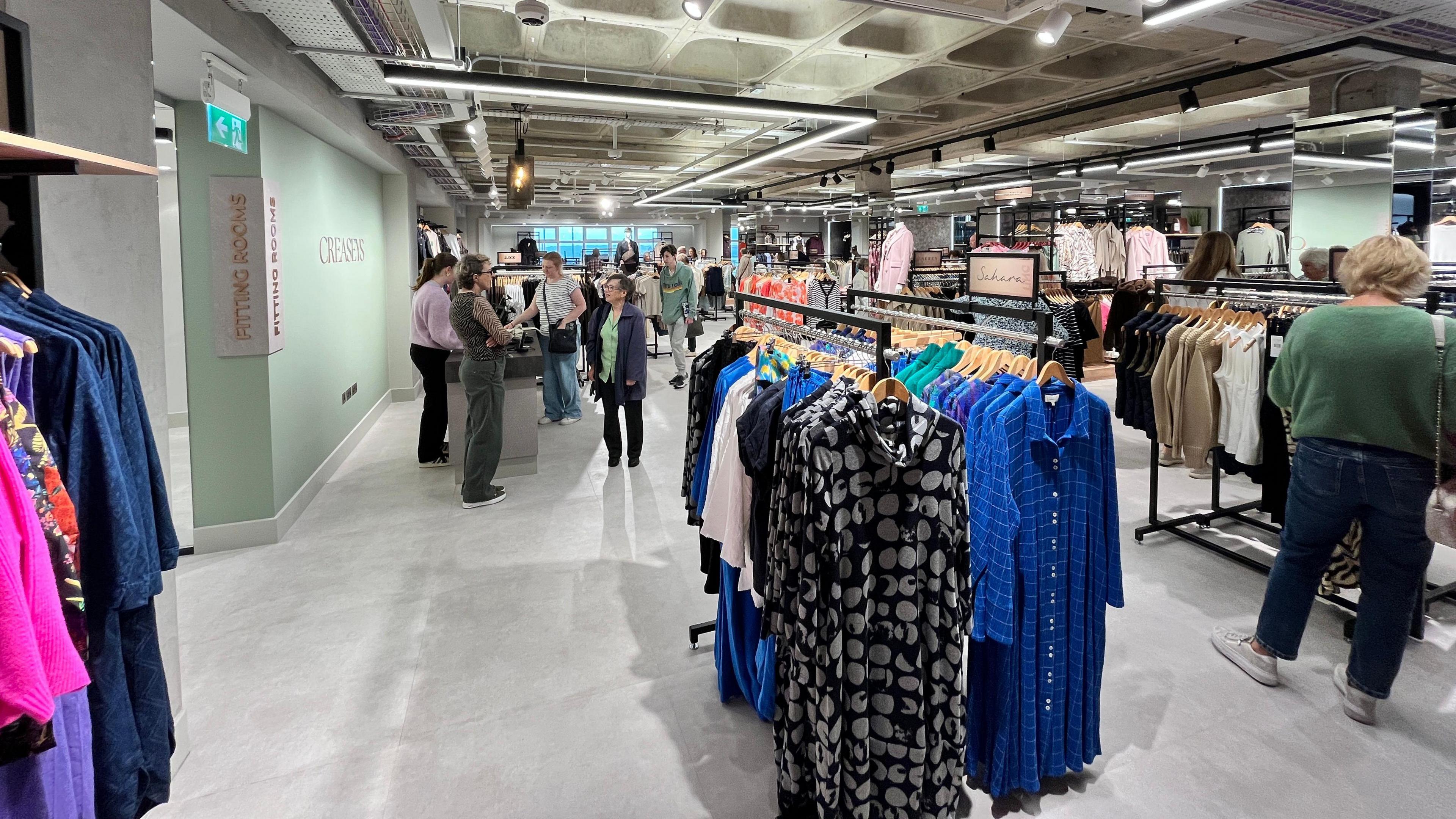 A large retail space with women's clothing including dresses on rails in a modern retail space. A mint green wall says Creaseys with a checkout next to it.