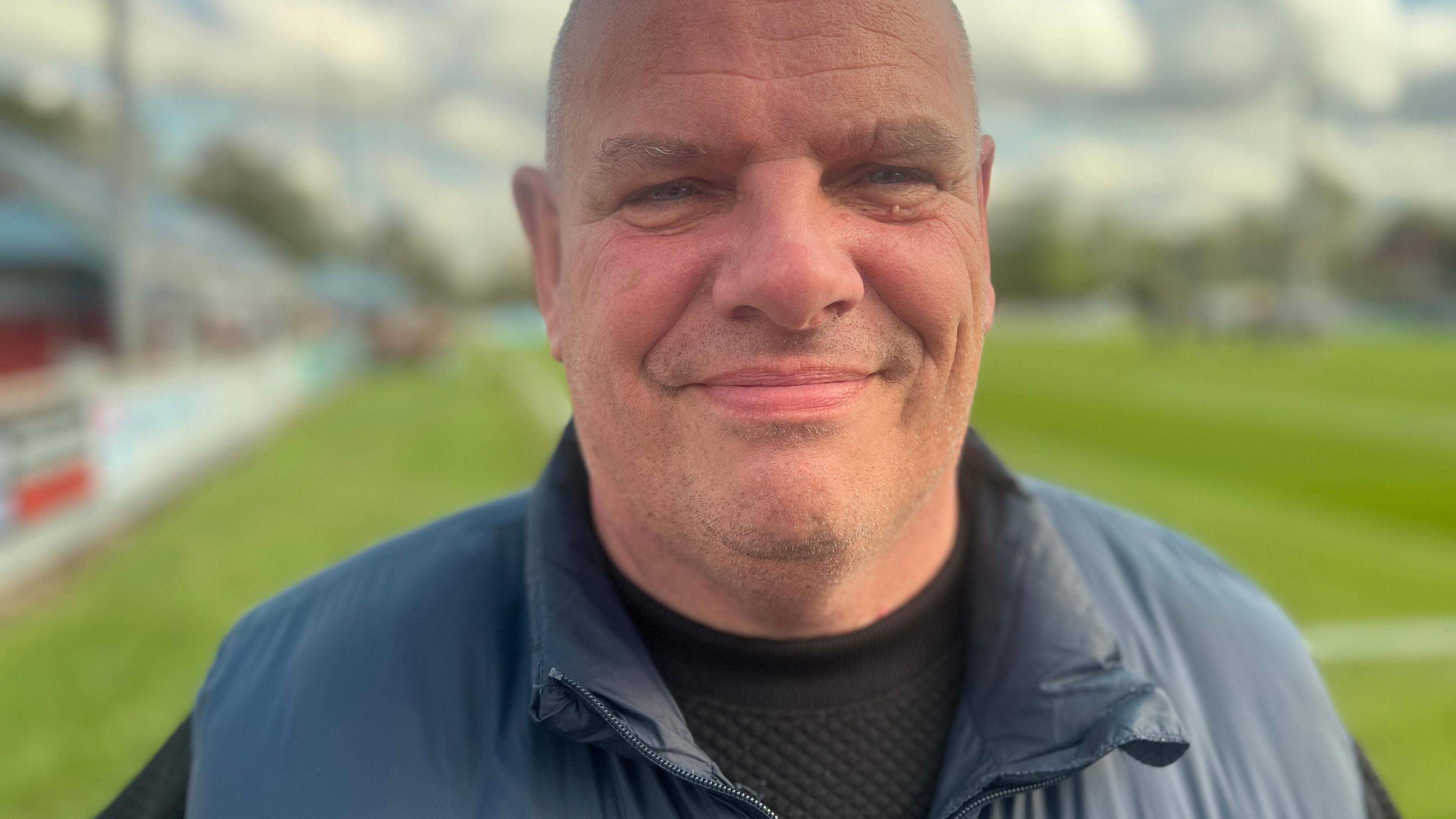 Karl Lindsay, in his 50s, who is bald and clean shaven, stands on a football pitch wearing a blue gilet over a black sweatshirt and smiles at the camera.