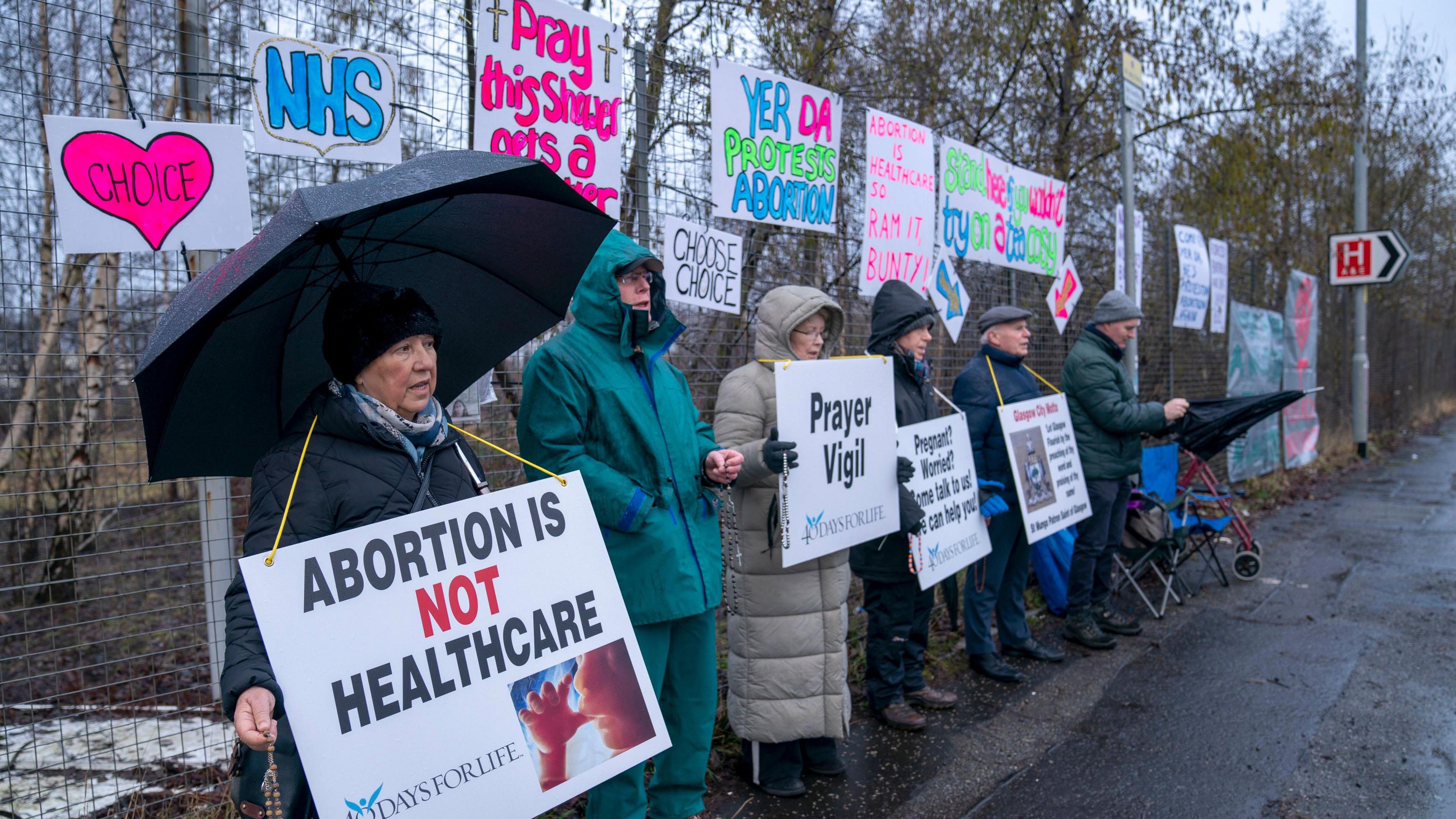 Six people in rainwear standing in a row, protesting against abortion. Their banners have slogans and photos of foetuses and have the 40 Days for Life branding.