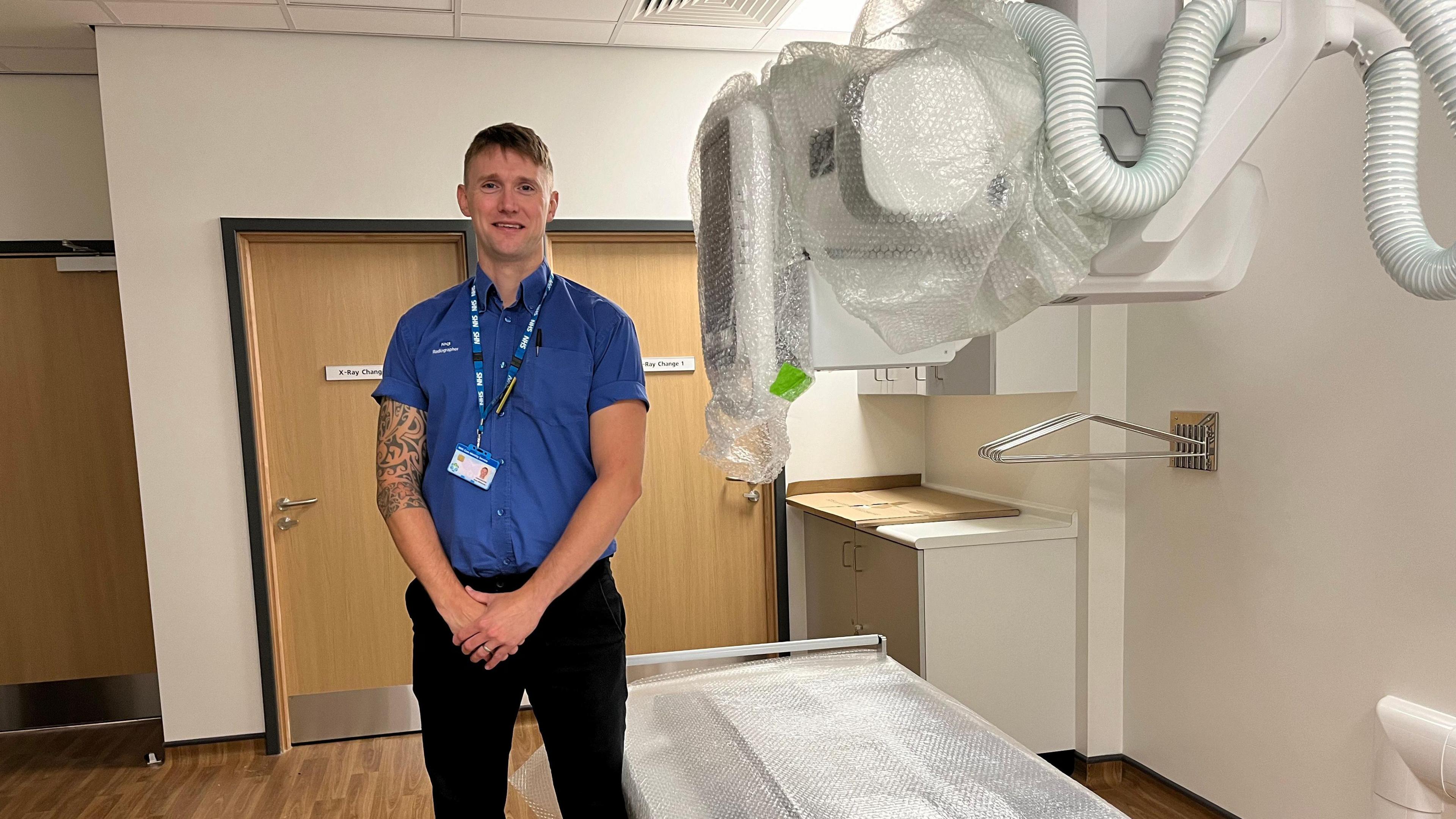 Alan Williams, clinical operations manager in radiology stands next to an x-ray machine which is so new the bubble wrap is still on it. He wears black trousers and a blue short sleeve shirt. He is tall and has tattoo's covering his right arm. He wears and NHS id card on a lanyard around his neck. 