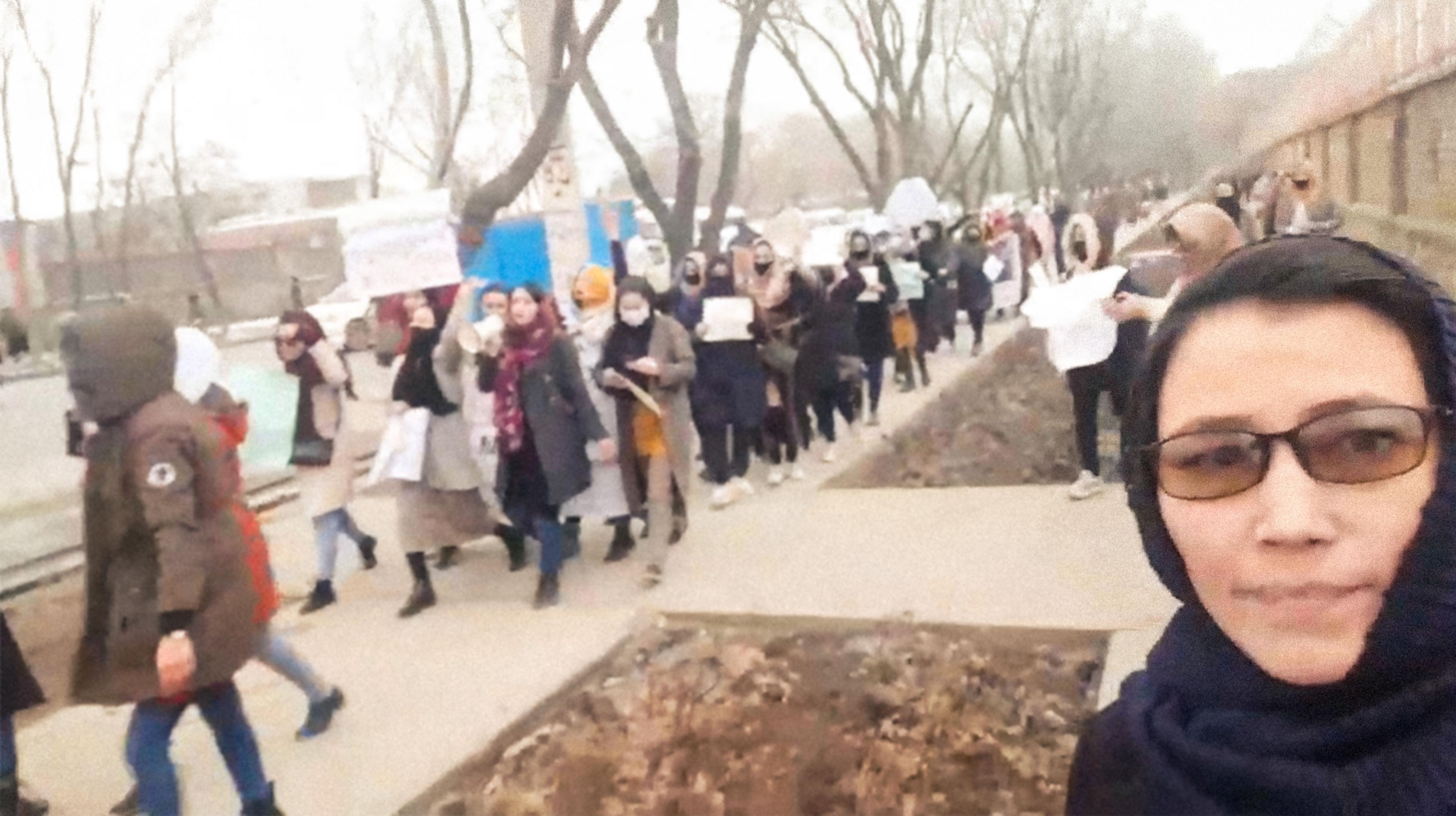 A still from Bread & Roses of a women's march. A long line of women walk along a path in Kabul holding placards while a woman dressed in a black dress and headscarf in the right-hand corner of the image takes a selfie. The city seems wintry, with no leaves on the trees and many of the protesters dressed in warm coats. 