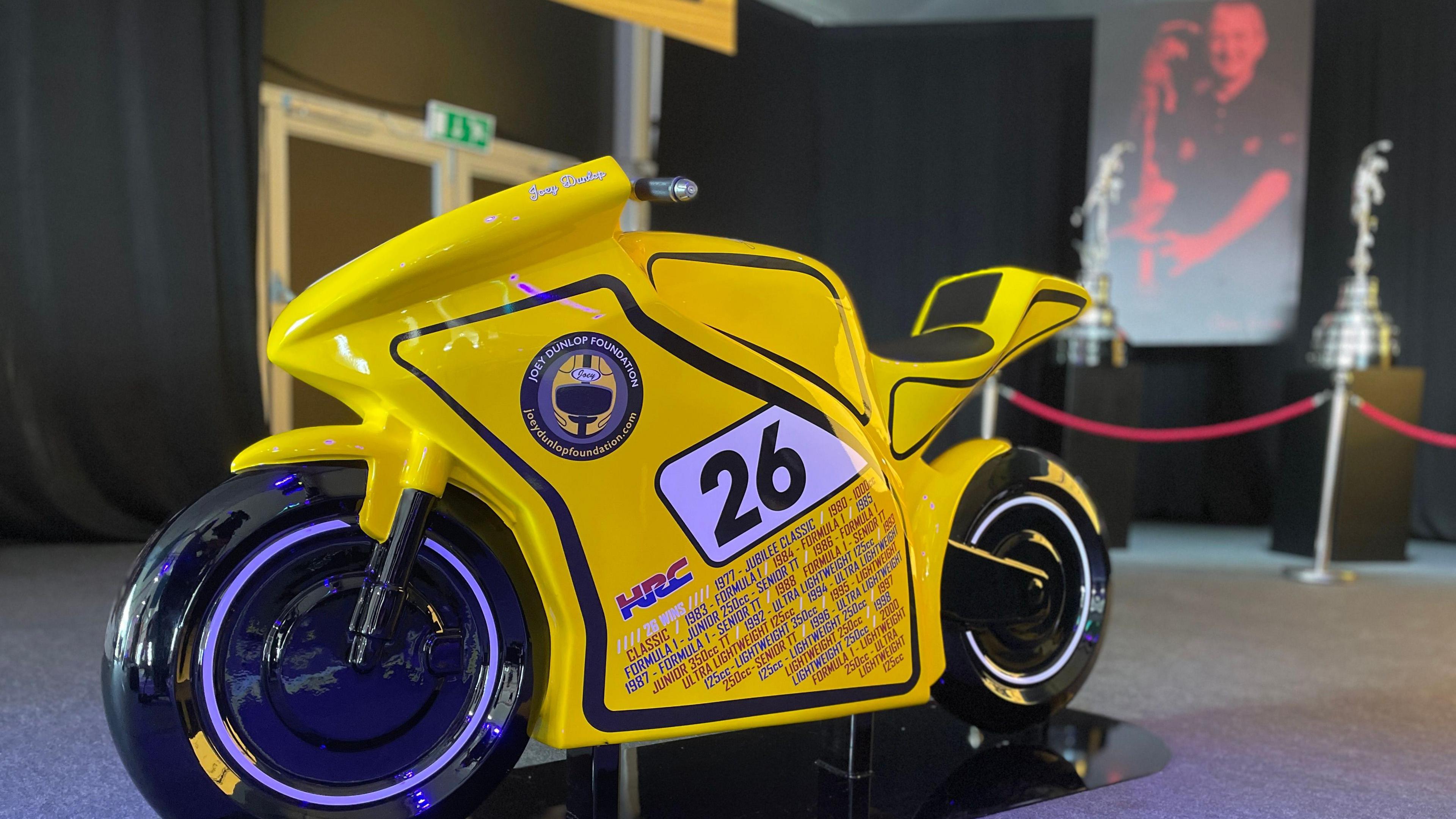 A life-size model yellow bike with the number 26 and text detailing all of Joey Dunlop's 26 wins.