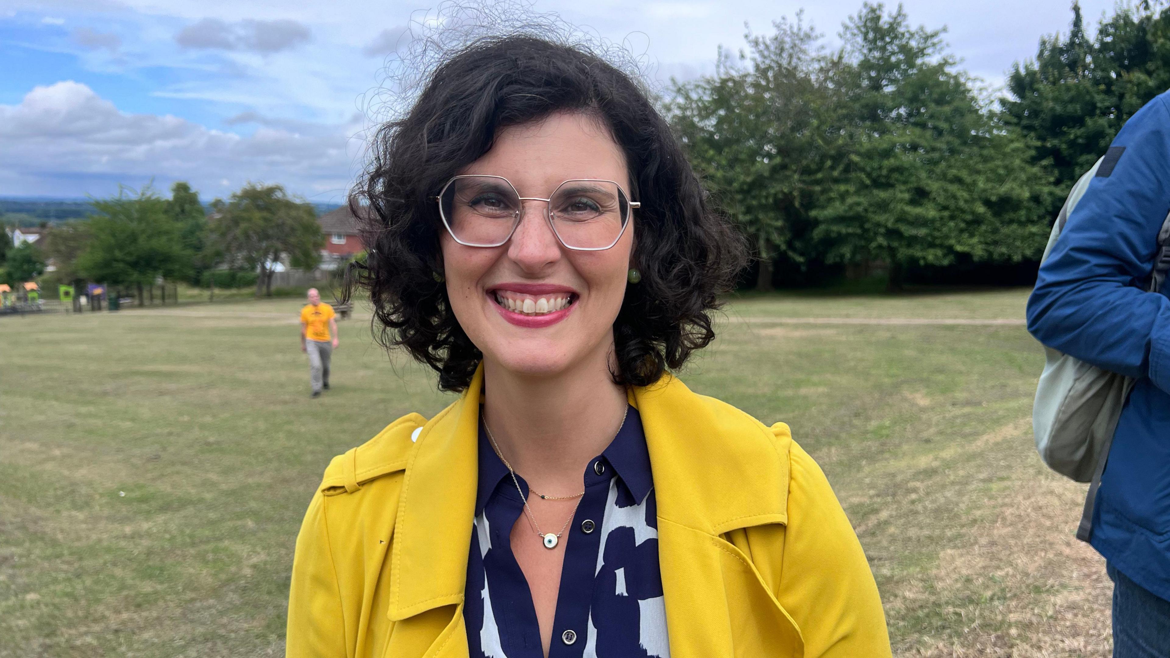 Layla Moran smiles at the camera, she has short curly black hair and wears large silver metal framed glasses with a blue and white print blouse and yellow trenchcoat over the top