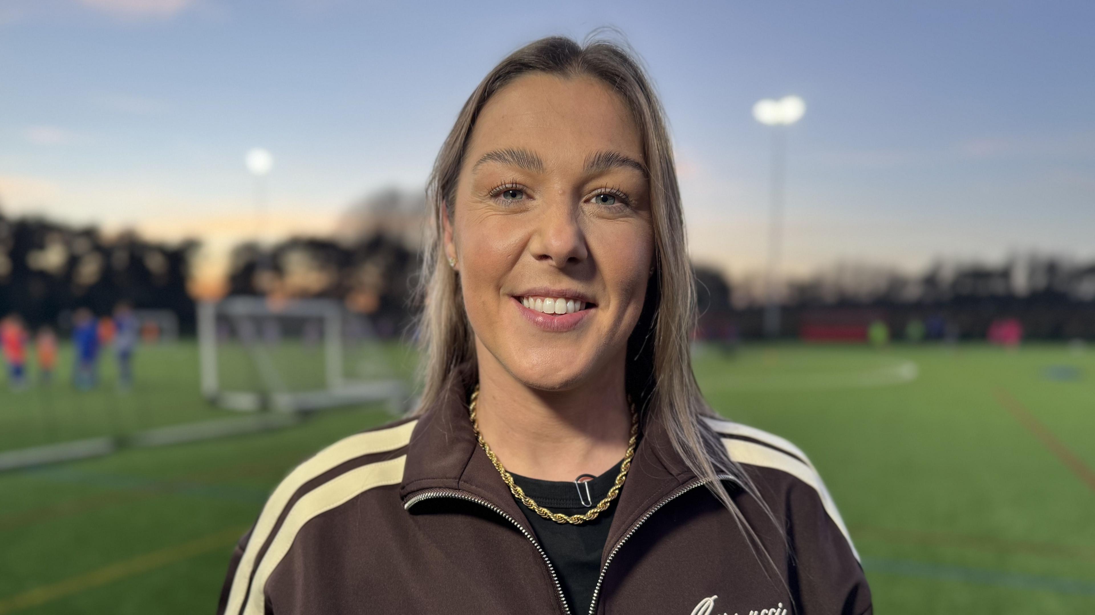 Mary Earps smiles as she looks into the camera and stands on the new football pitch. She's wearing a black top underneath a brown jacket with cream stripes down the shoulders as well as a gold necklace.