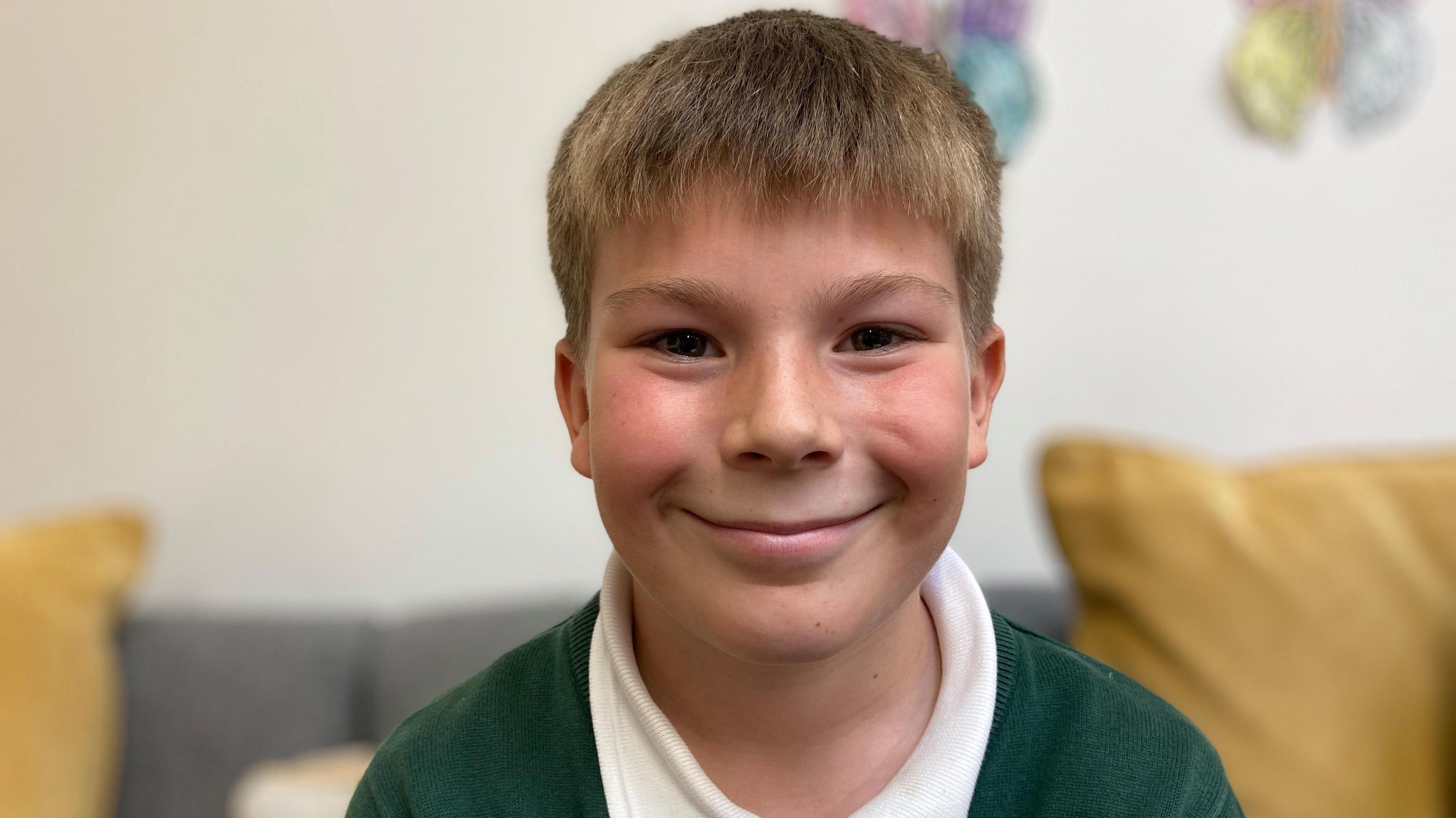 A smiling Ethan looks directly at the camera as he is photographed wearing his green school uniform. He has short blond hair and is sitting on a grey sofa.