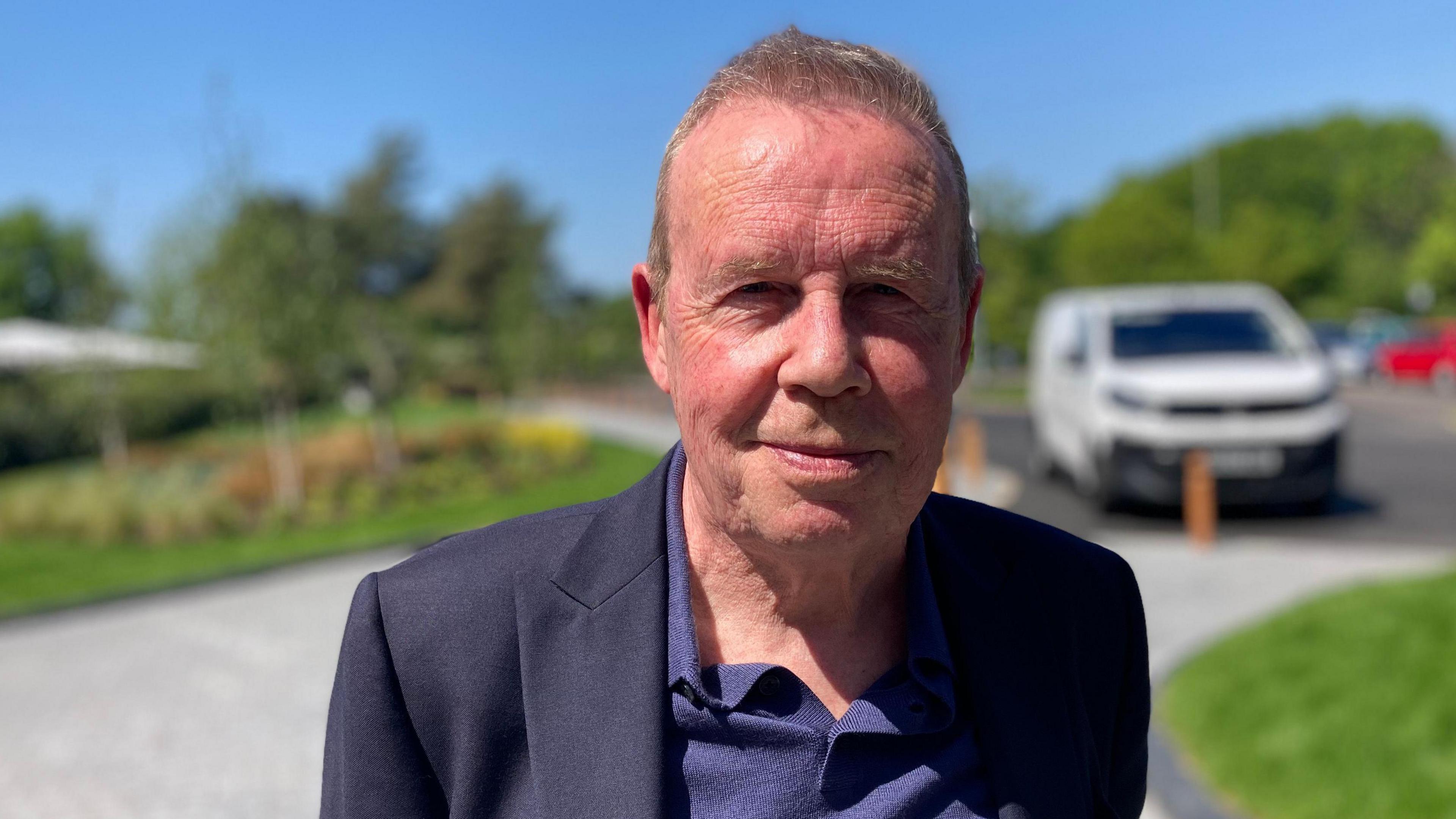 A man in his seventies wearing a dark shirt and jacket with a ruddy complexion.