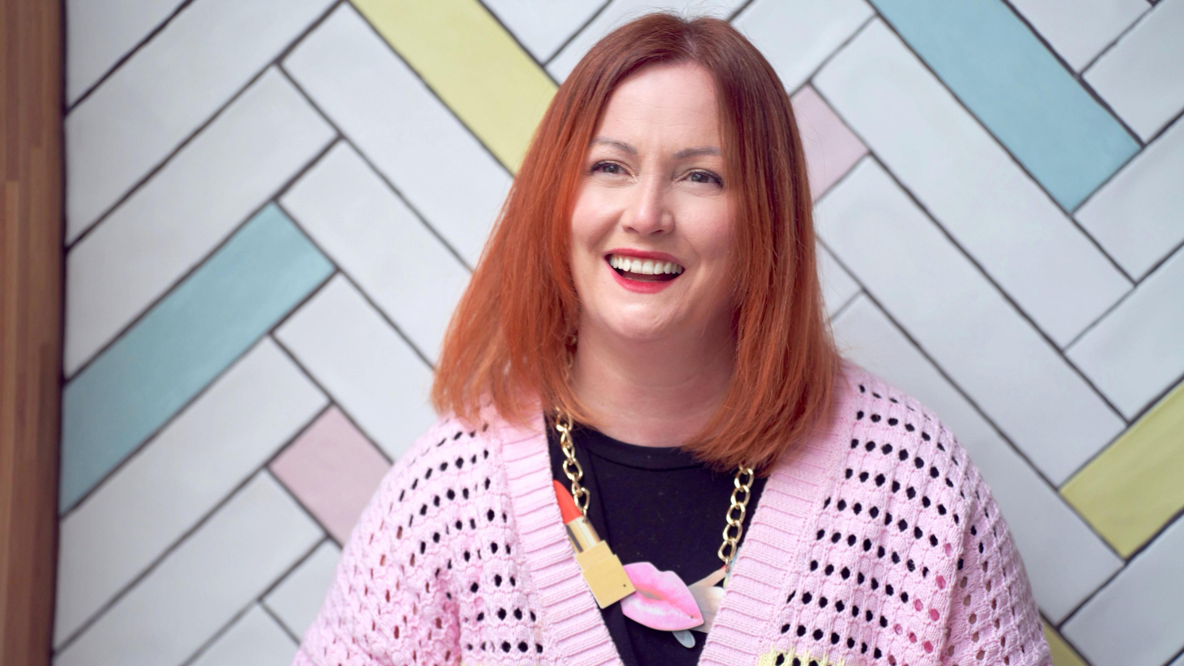 Dr Sedgman stands in front of pastel-coloured tiles. She has shoulder length copper hair and smiles at the camera. She wears a pink cardigan, with a black top underneath and a chunky necklace. 