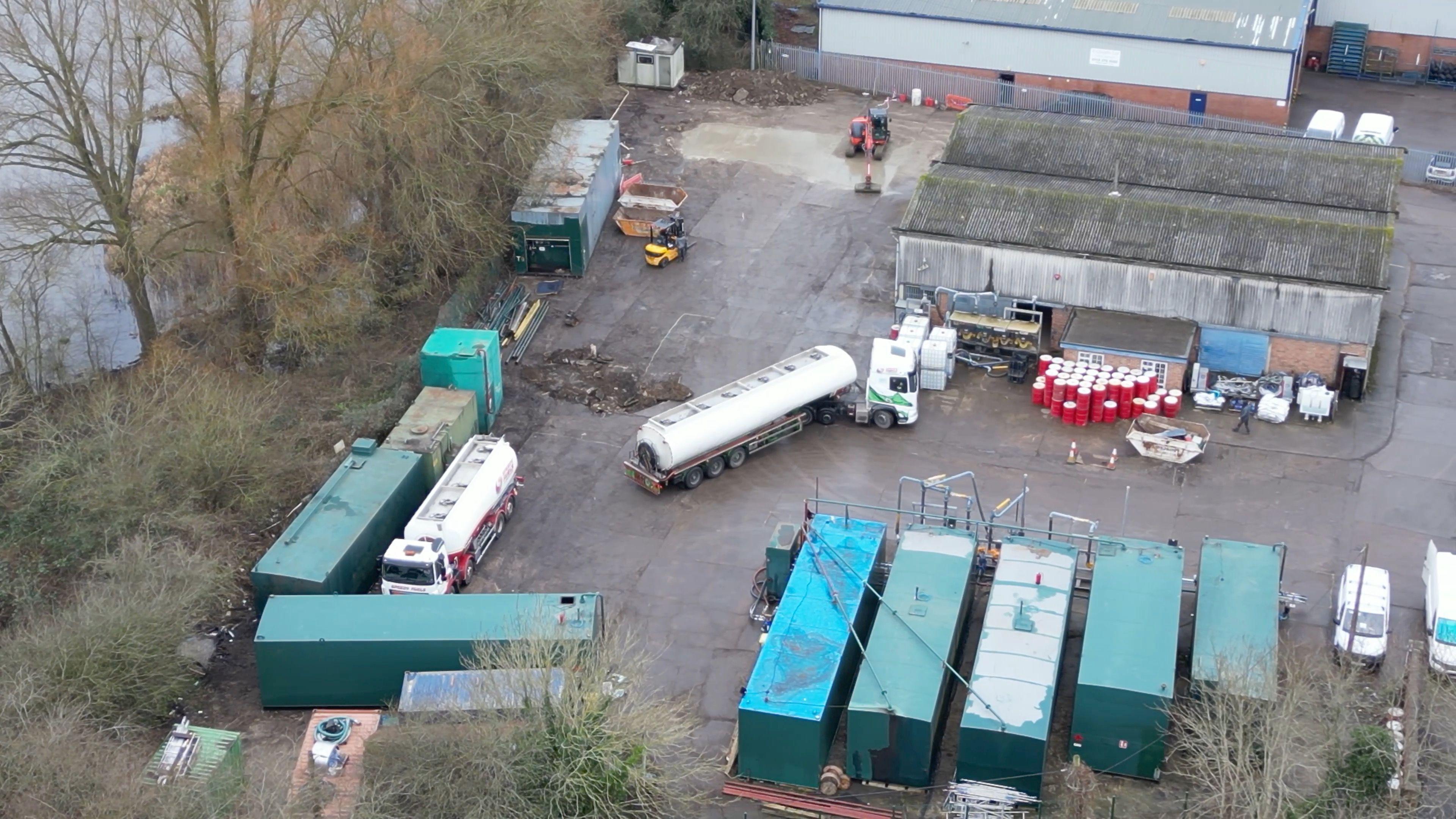 A drone photo showing oil tankers moving around a concrete yard. There's some trees and the edge of a lake directly next to it.