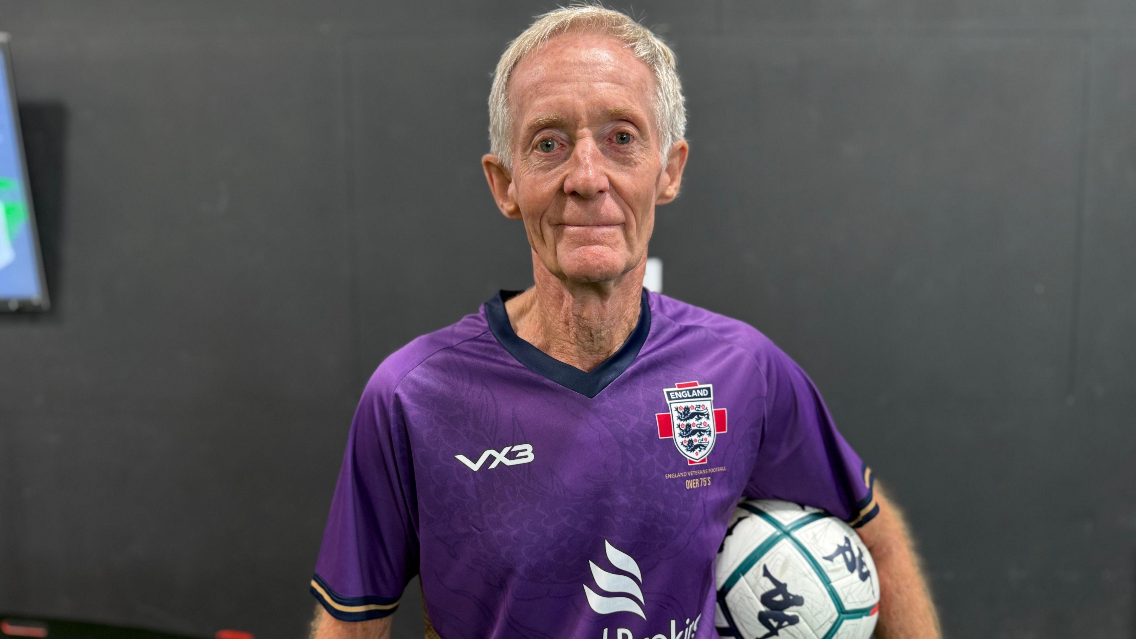 Photo of a man with short grey hair looking into the camera. He is wearing a purple football shirt with an England badge on the right. He is also holding a while football with turquoise detailing.