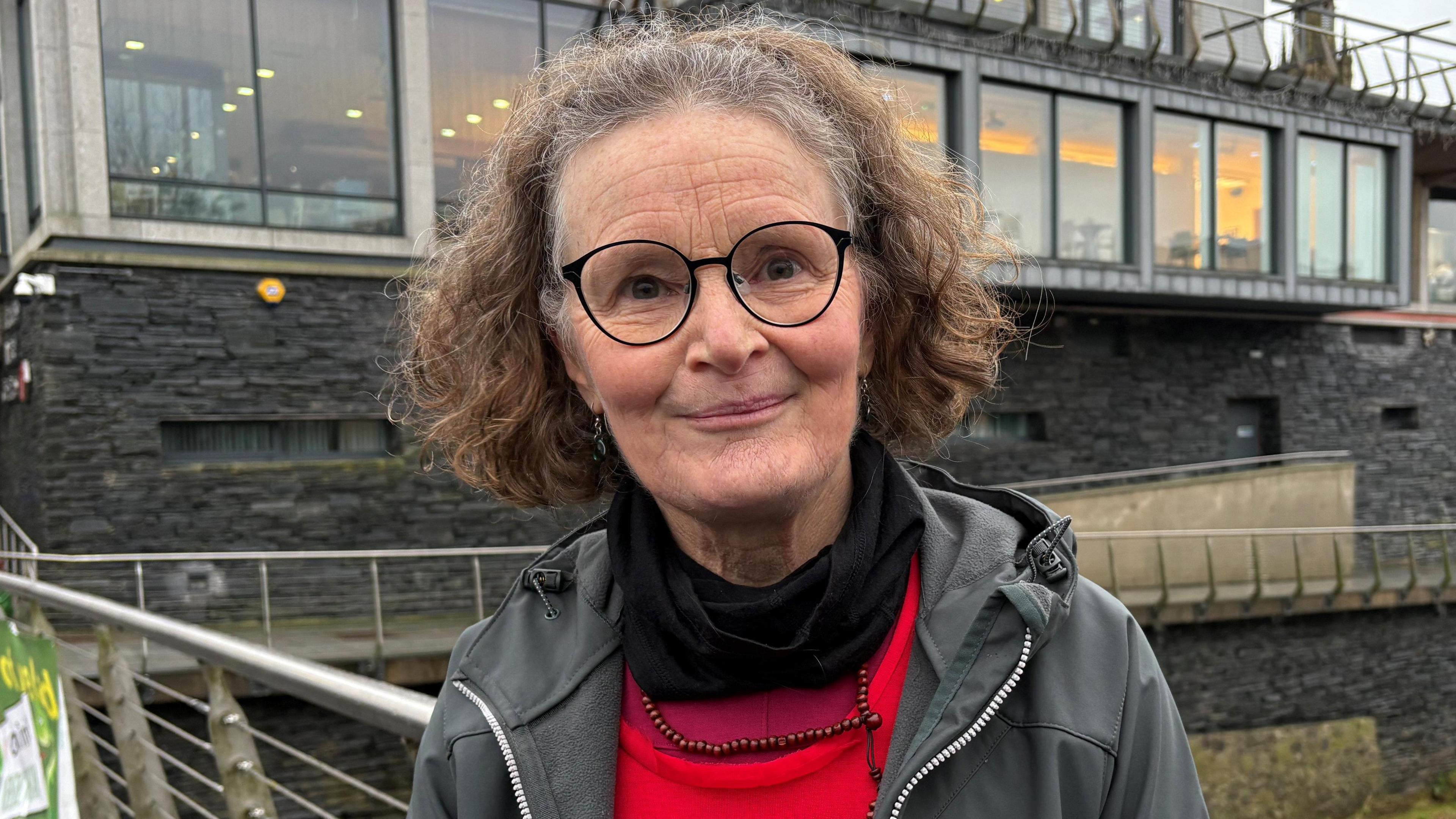 Marella is smiling into the camera. She's standing on a bridge with a building behind her. She has short brown hair and is wearing a red top to show that she's protesting and a grey hoodie.