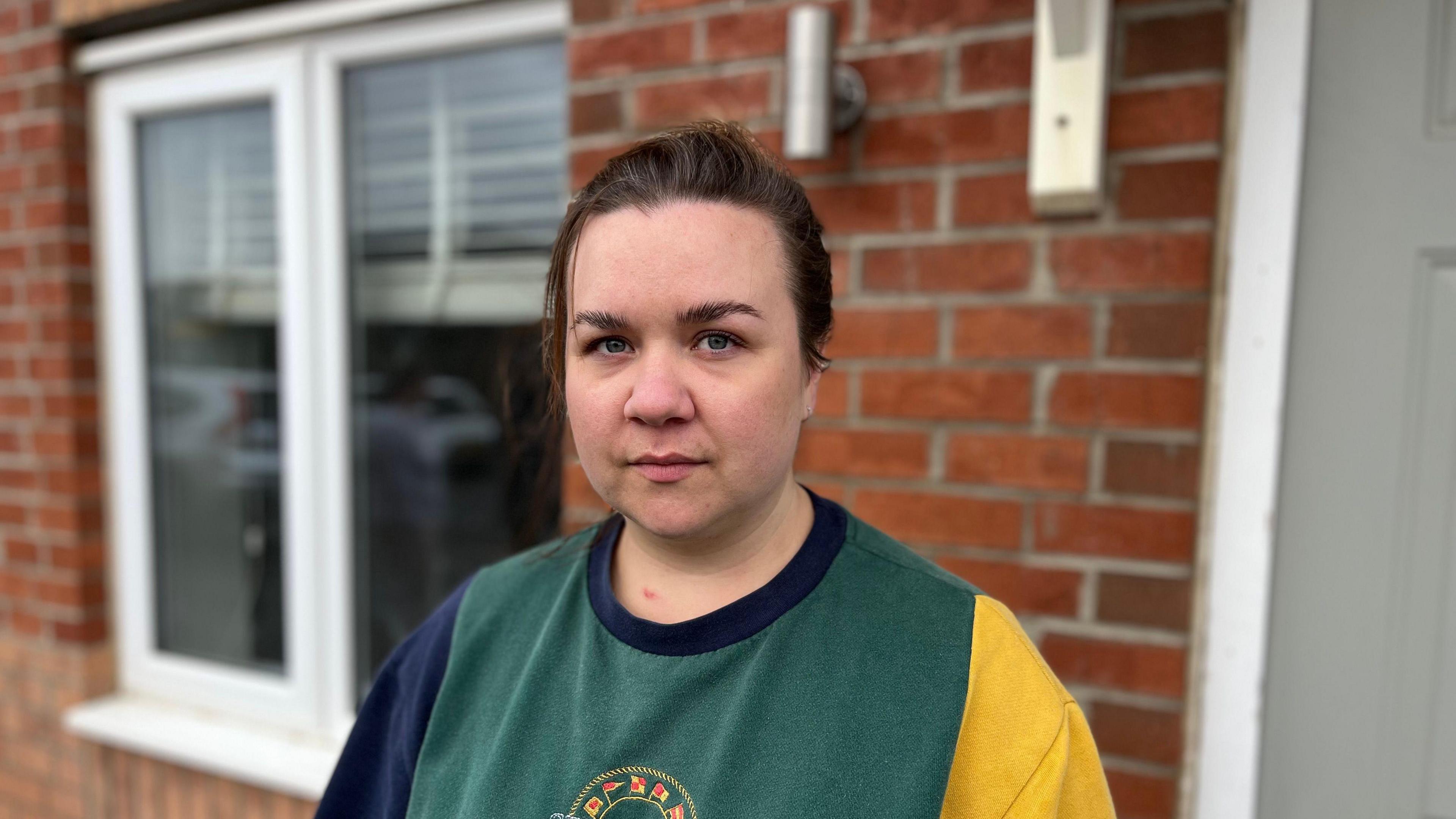 Kerry Forster has brown hair tied back into a ponytail. She is standing in front of her house and looking into the camera. She is wearing a green jumper.