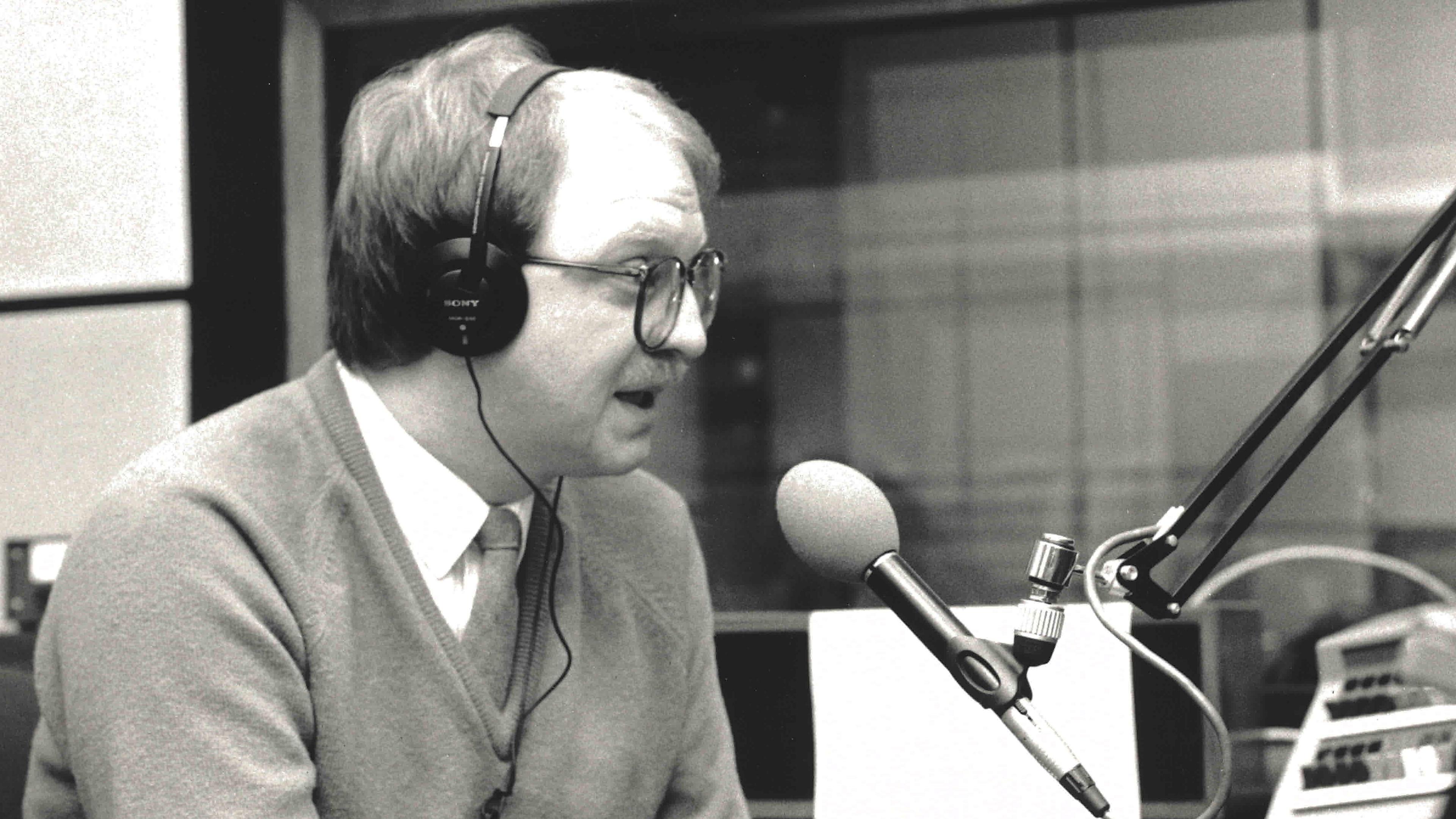 Gavin McCoy is wearing a V-neck jumper over a white shirt and tie. He is speaking into a microphone while wearing headphones.