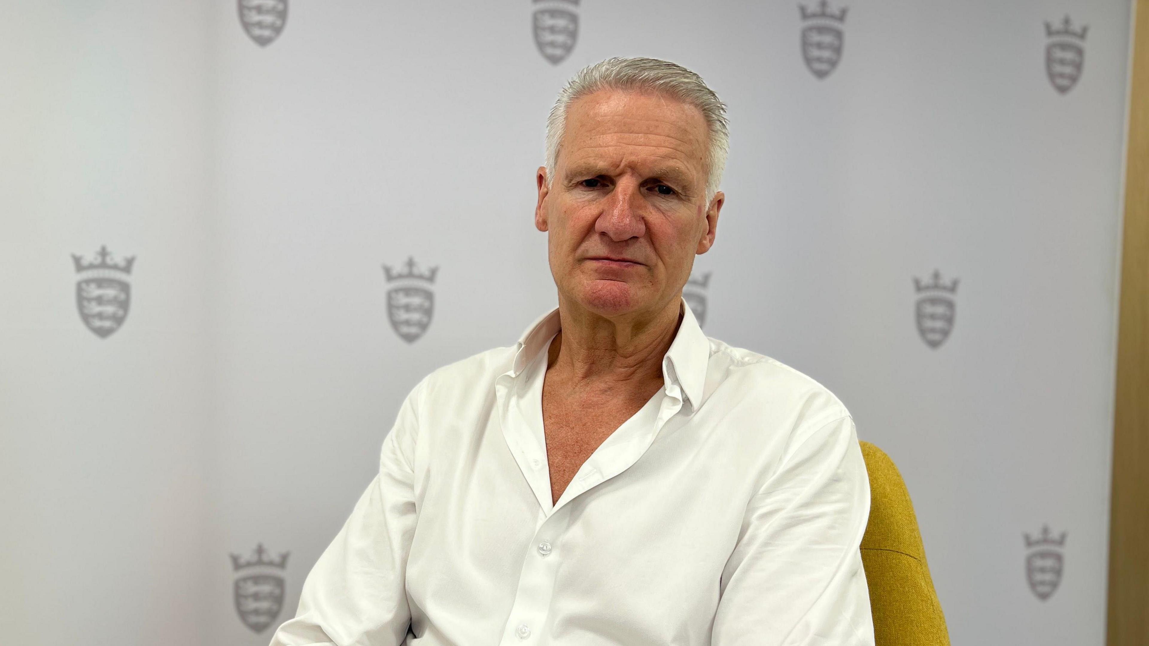 Jersey Health and Social Services Minister Tom Binet sits on a yellow chair in front of a board with the Government of Jersey's logo on it. He is wearing a white shirt with the top couple of buttons undone.