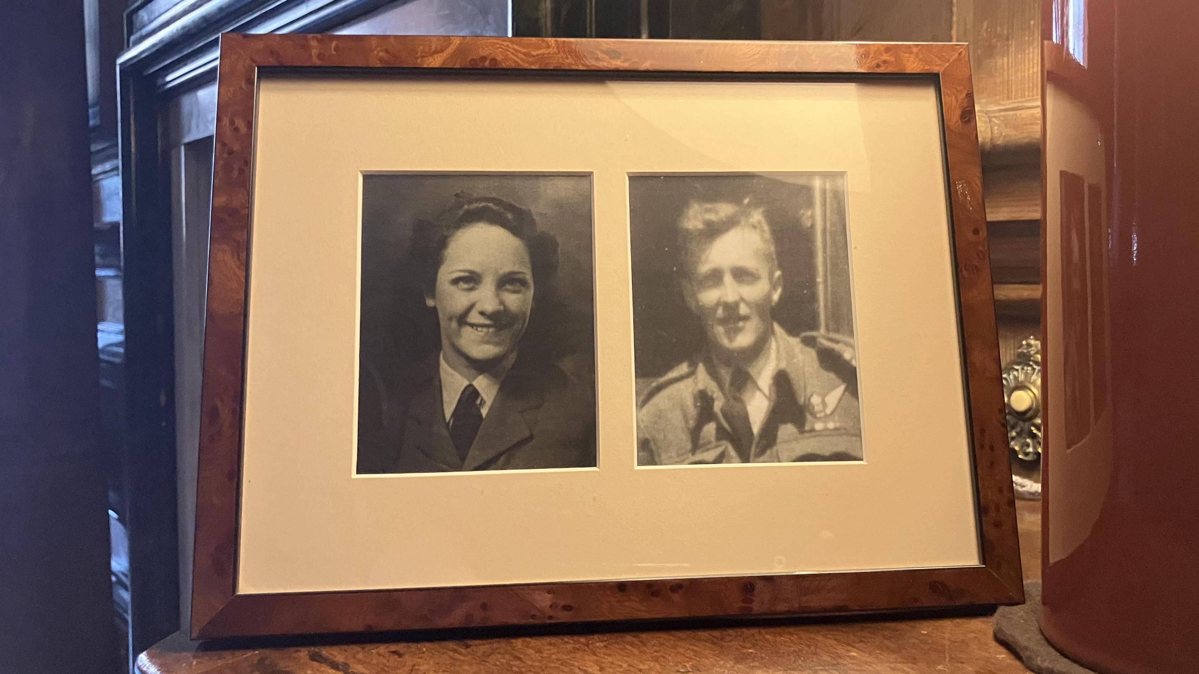 A photo frame with black and white headshots of a woman on the left hand side and a man on the right, in a dark oak frame.