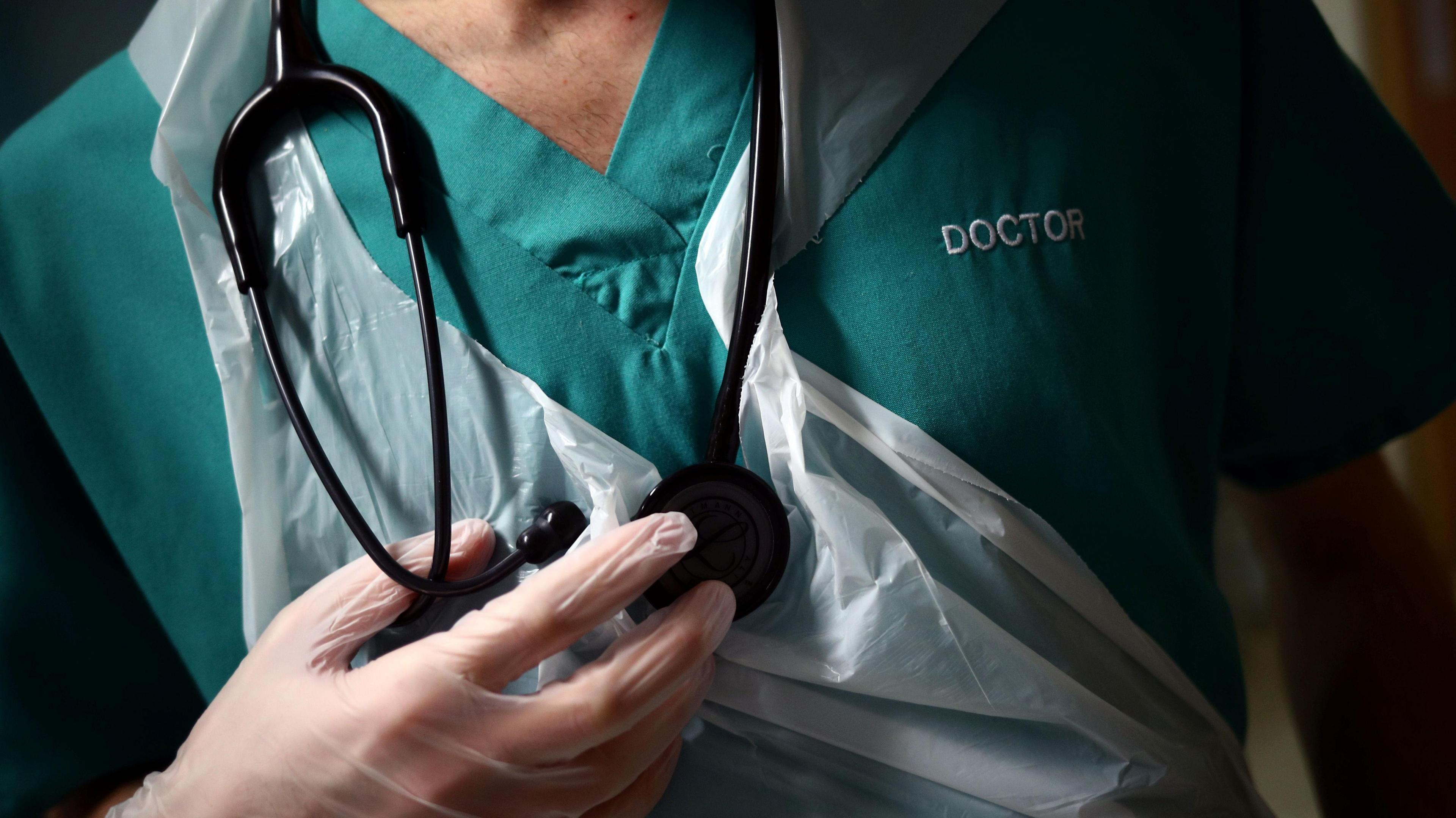 Close-up a headless doctor holding a stethoscope. He is wearing a green v-neck overall top with DOCTOR embroidered on he left hand side in white. He is also wearing an apron and a pair of latex gloves, but only his right hand is visible in the shot.