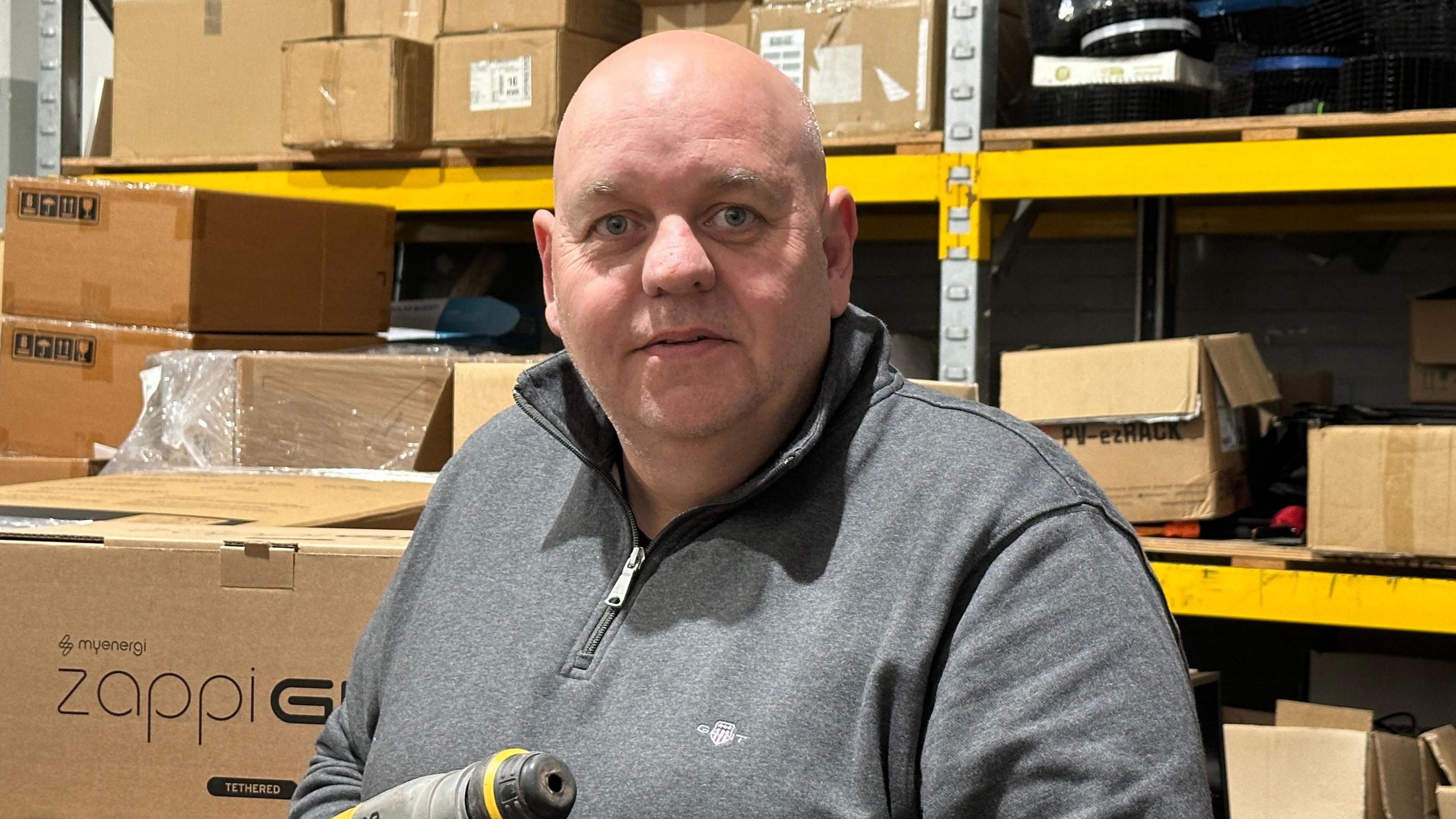 Wayne Brightmore in a warehouse with boxes stacked on shelves behind him. He is bald and is wearing a grey quarter-zip. He is looking at the camera and holding a screwdriver. 