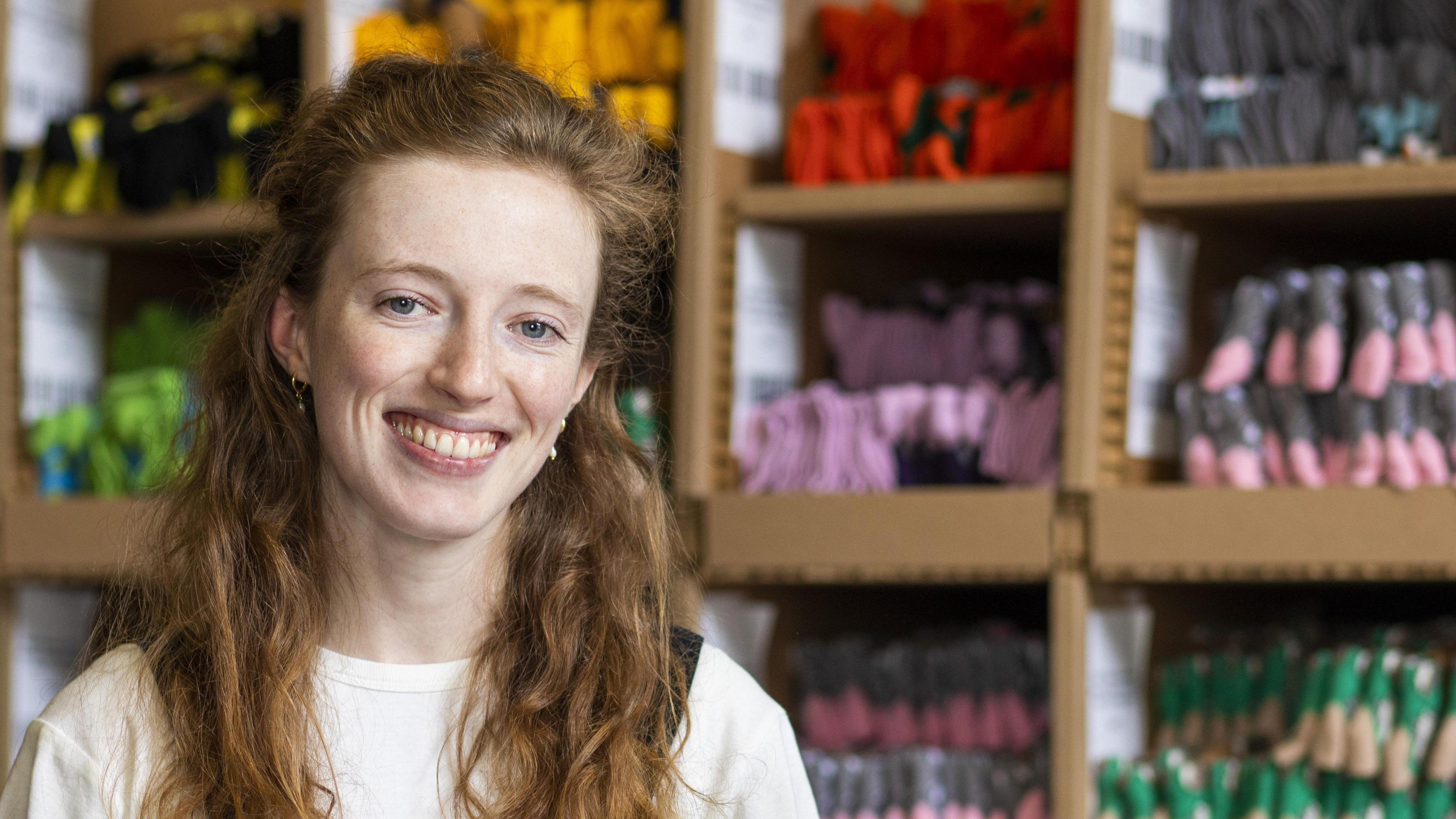 A lady with long red hair wearing a white jumper is sat in front of shelves which have lots of socks neatly folded in them.