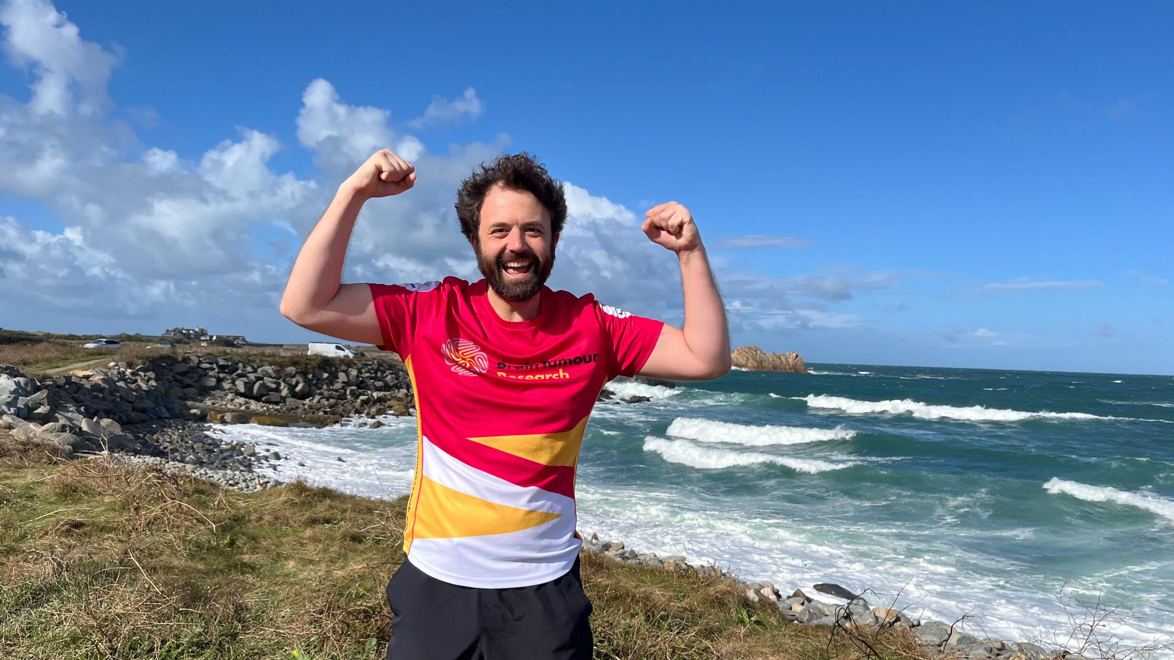 A man in a red, white and yellow top that says 'Brain Tumour Research'. He's got dark curly hair and a beard and has his arms in a 'strong' position, with his fists clenched. Behind him is the sea, which is a dark blue colour. The sky is blue with white clouds.