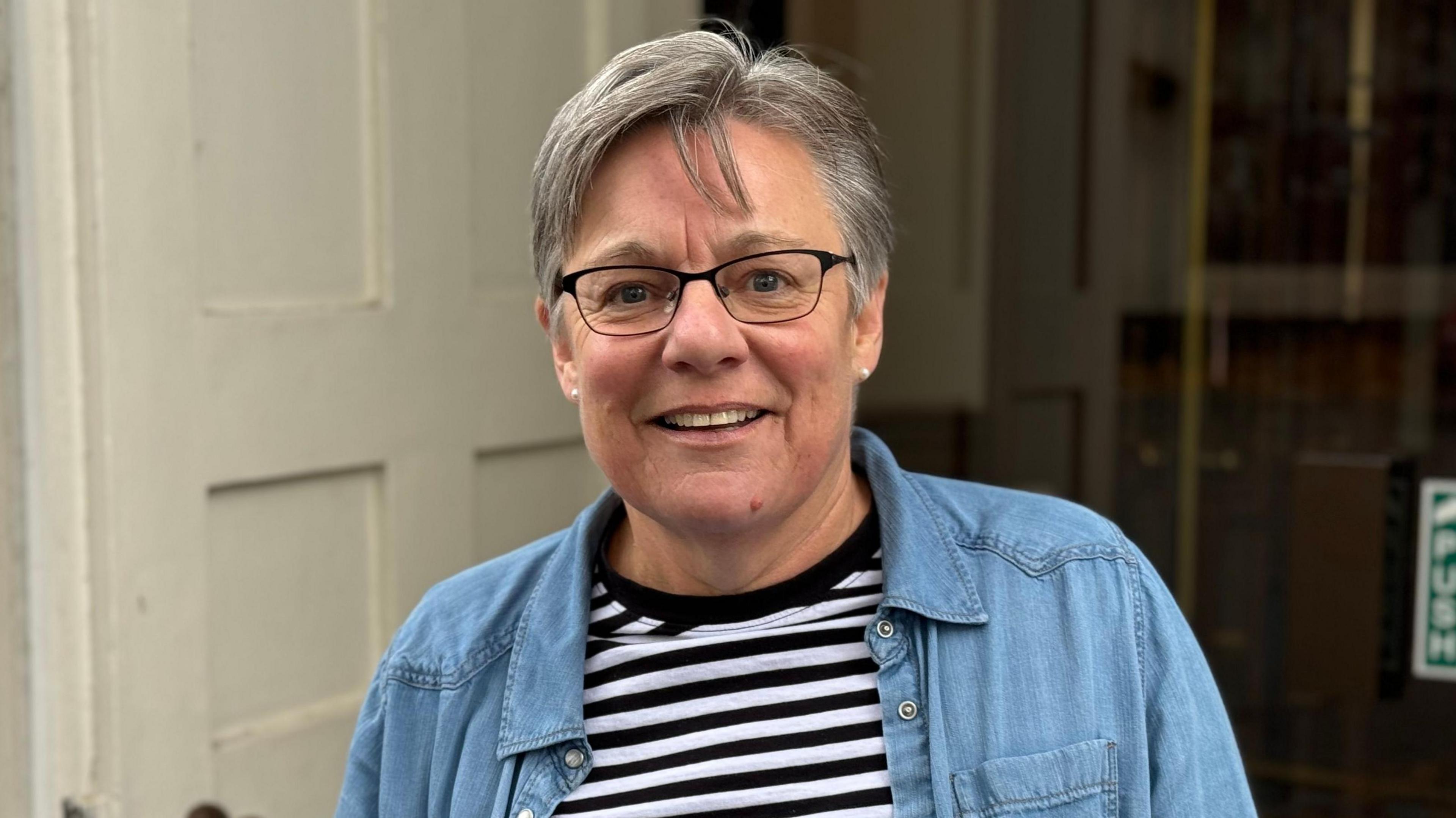 A woman with black rimmed glasses and grey hair looking at the camera. She is wearing a blue denim overshirt and a striped black and white shirt. 