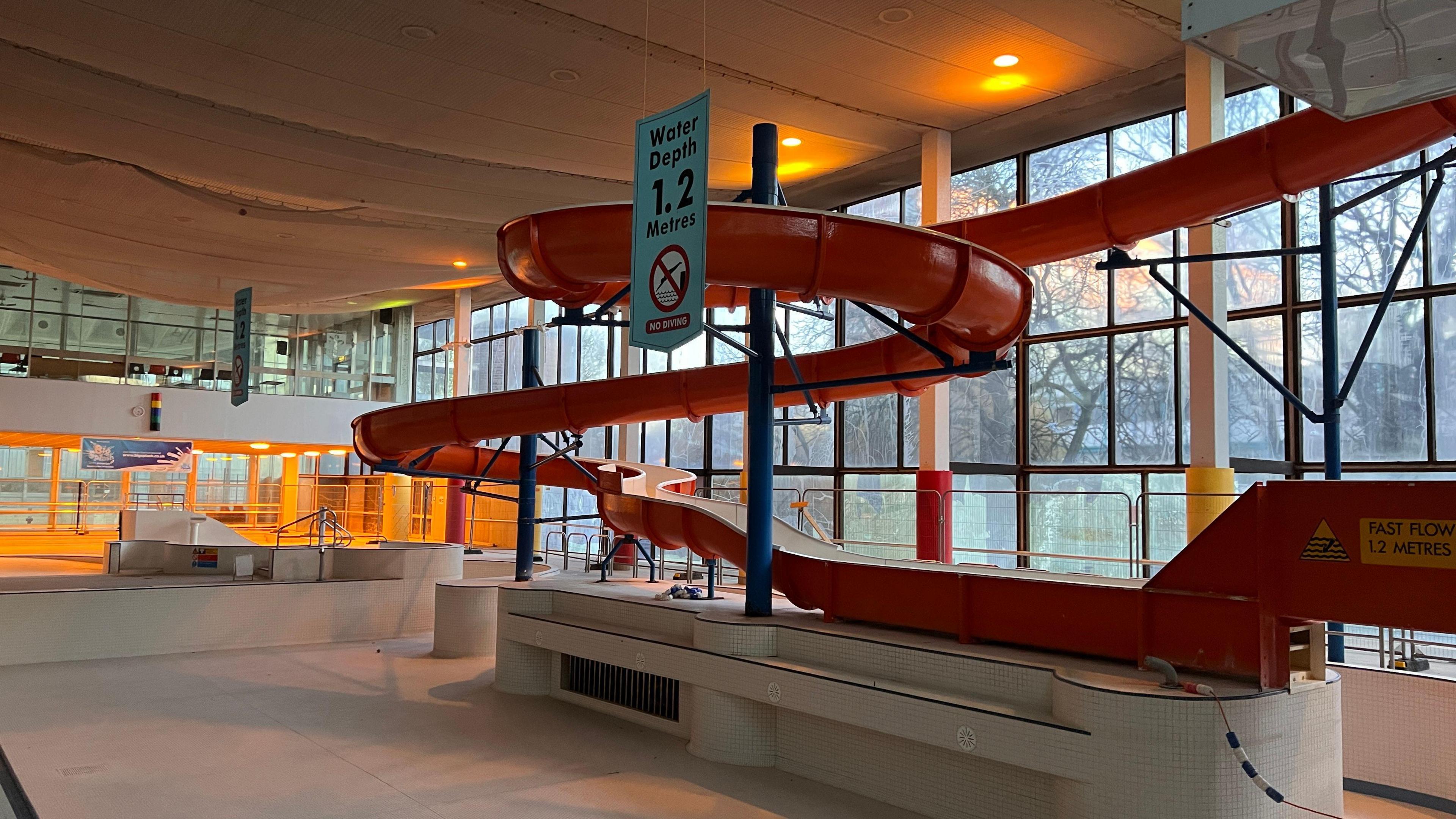 An abandoned red water slide in the swimming pool building with the pool surrounding it drained of any water.
