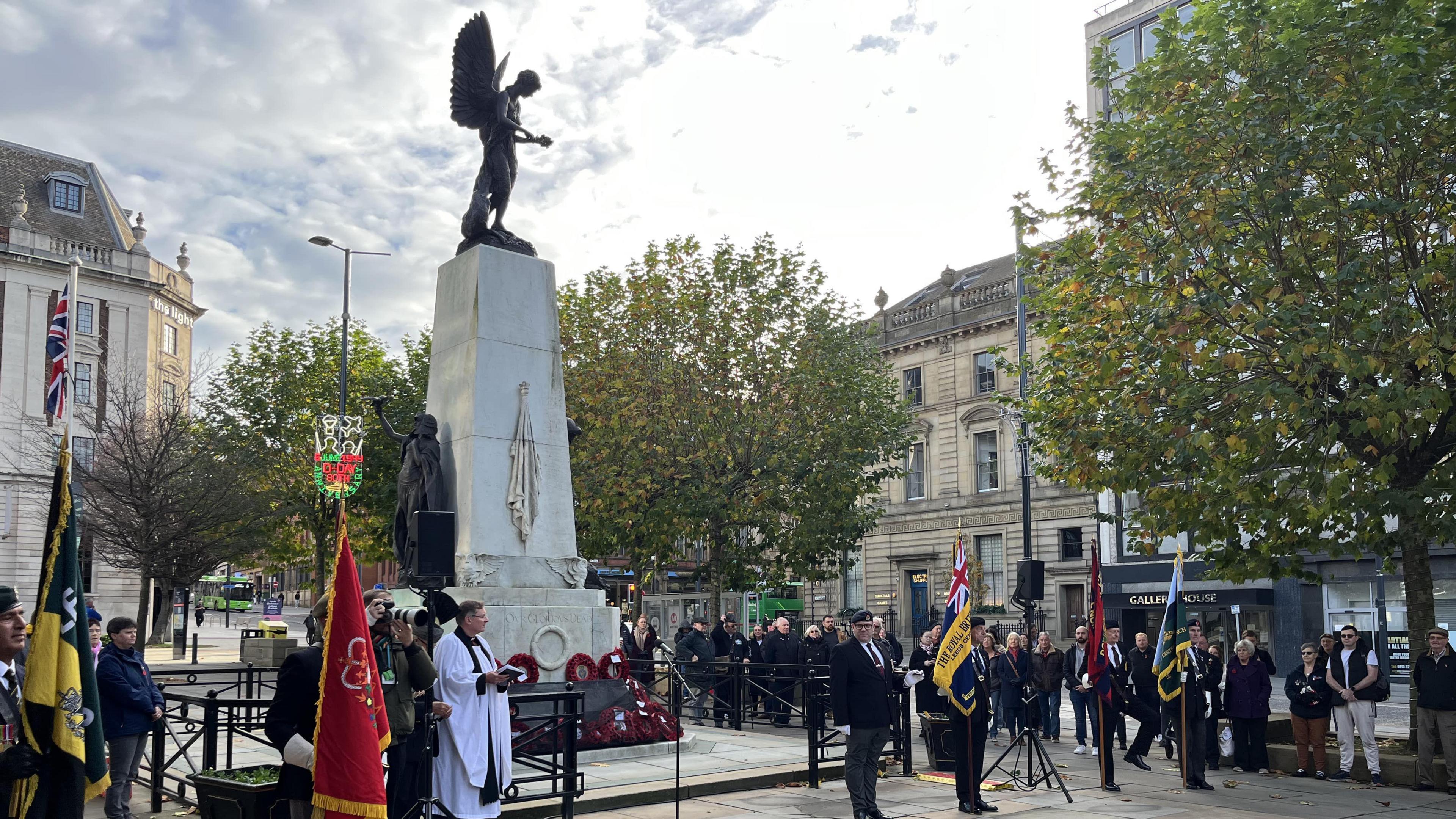 An Armistice Day service is held at the War Memorial in Leeds. Military flags are held aloft by service personnel as a vicar stands at the front of the monument. Crowds line the side.