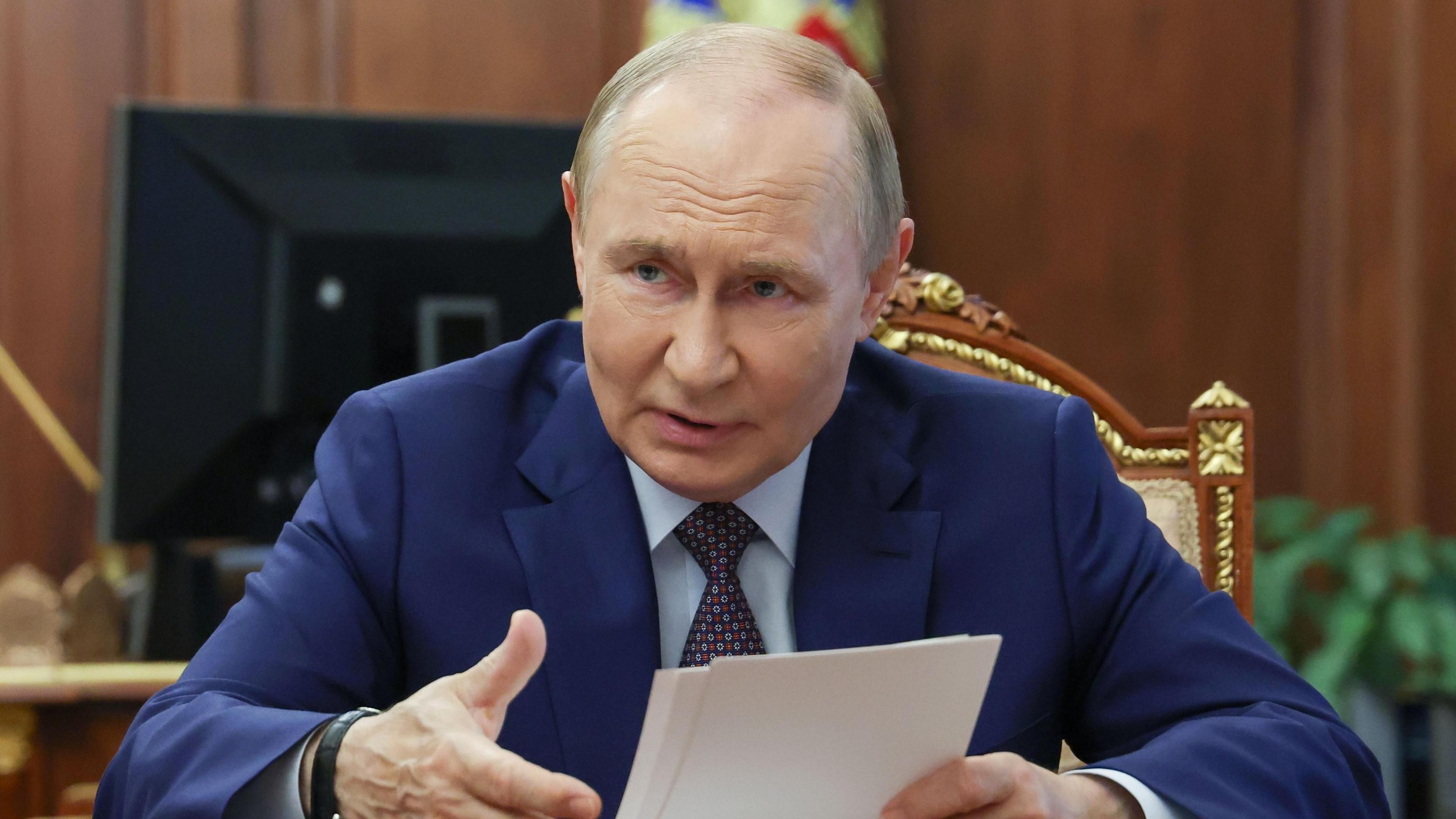 A balding man is wearing a dark blue suit, with a light blue shirt and tie. He is gesturing at some papers in his hand.