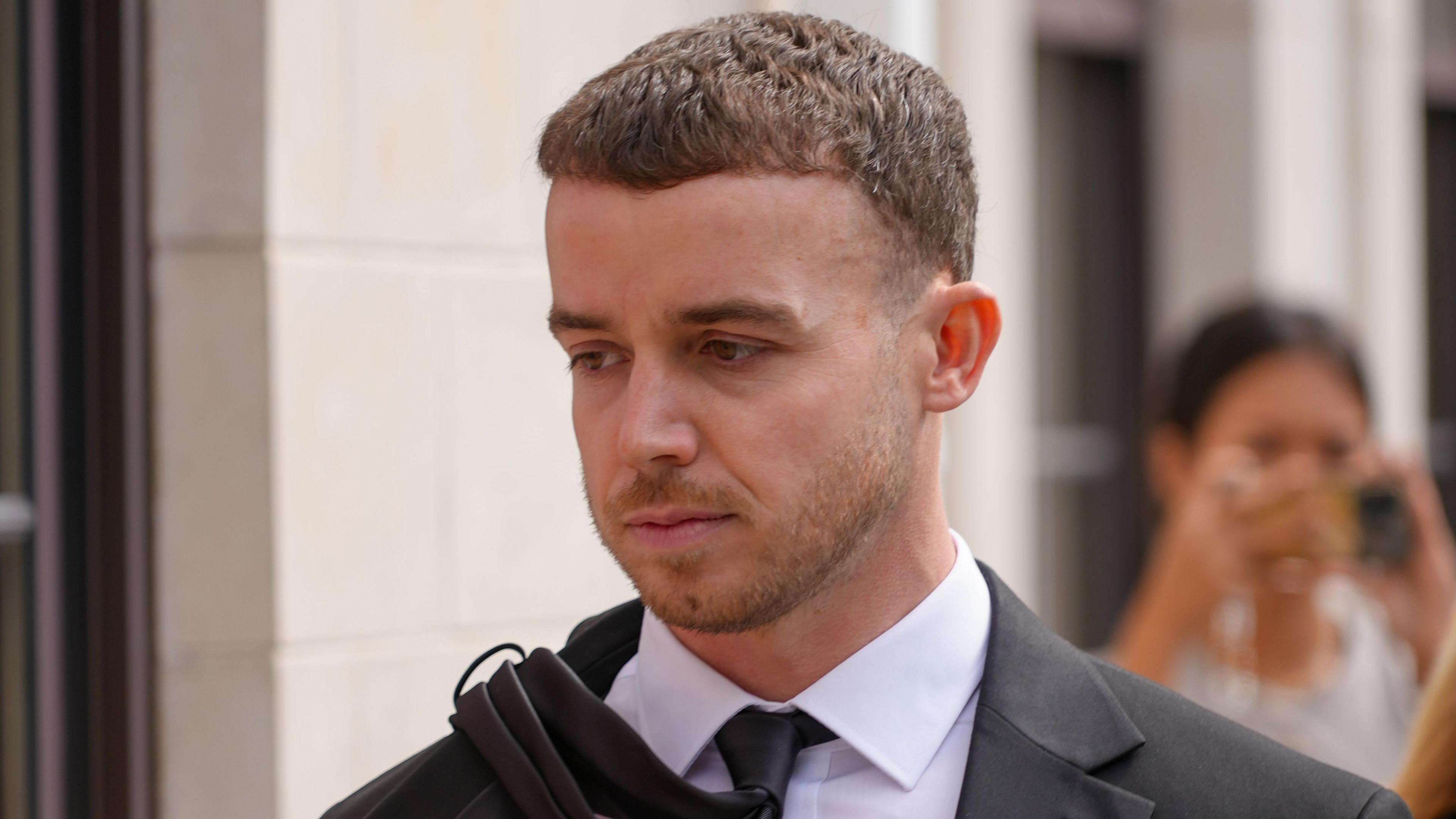 A man wearing a suit and tie walking into court, background blurred but woman can be seen taking a photo.