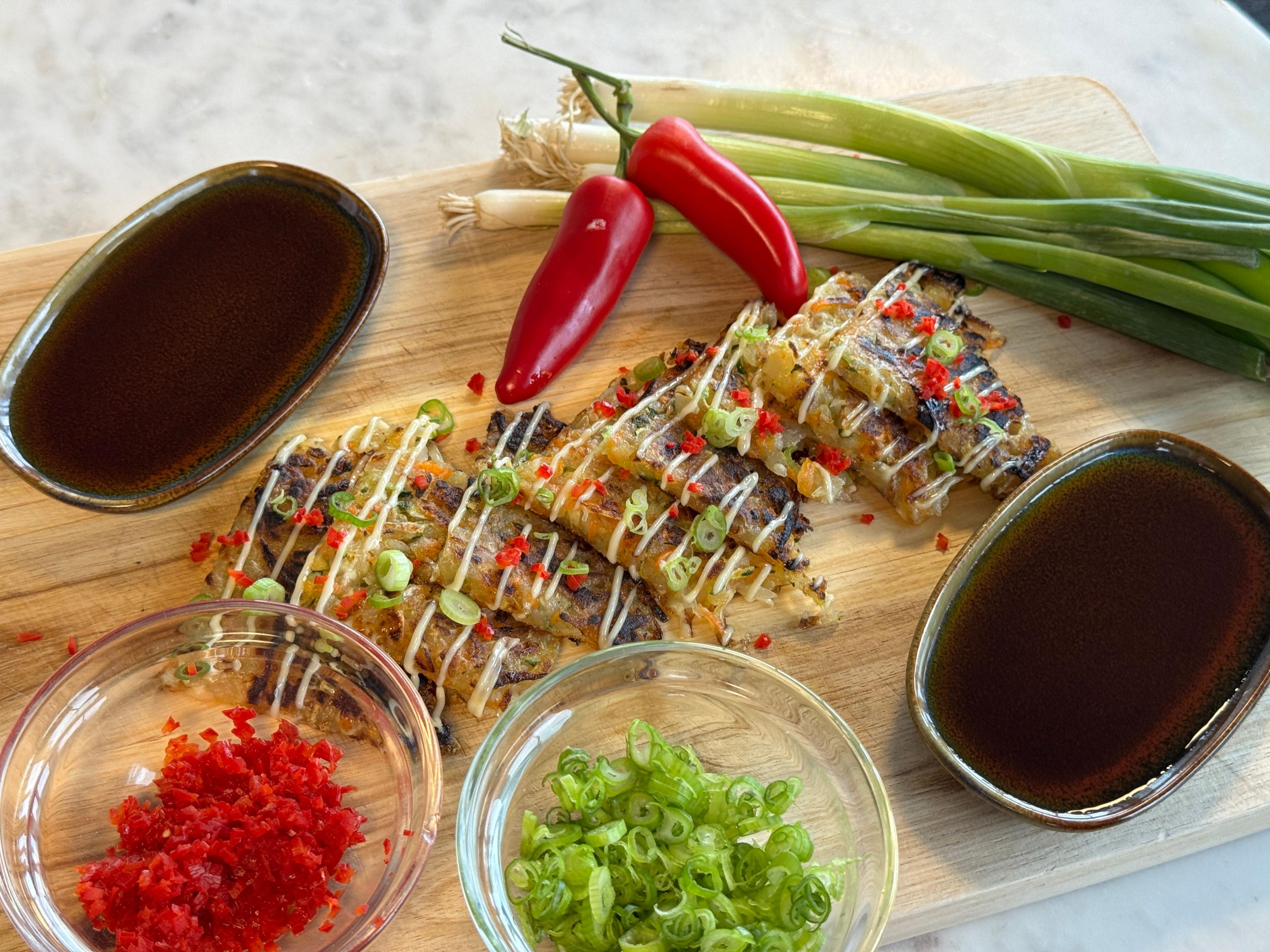 Crispy pancakes in the middle of a wooden chopping board, drizzles in a mayonnaise, surrounded by pots of chilli's, green onions, and soy sauce.