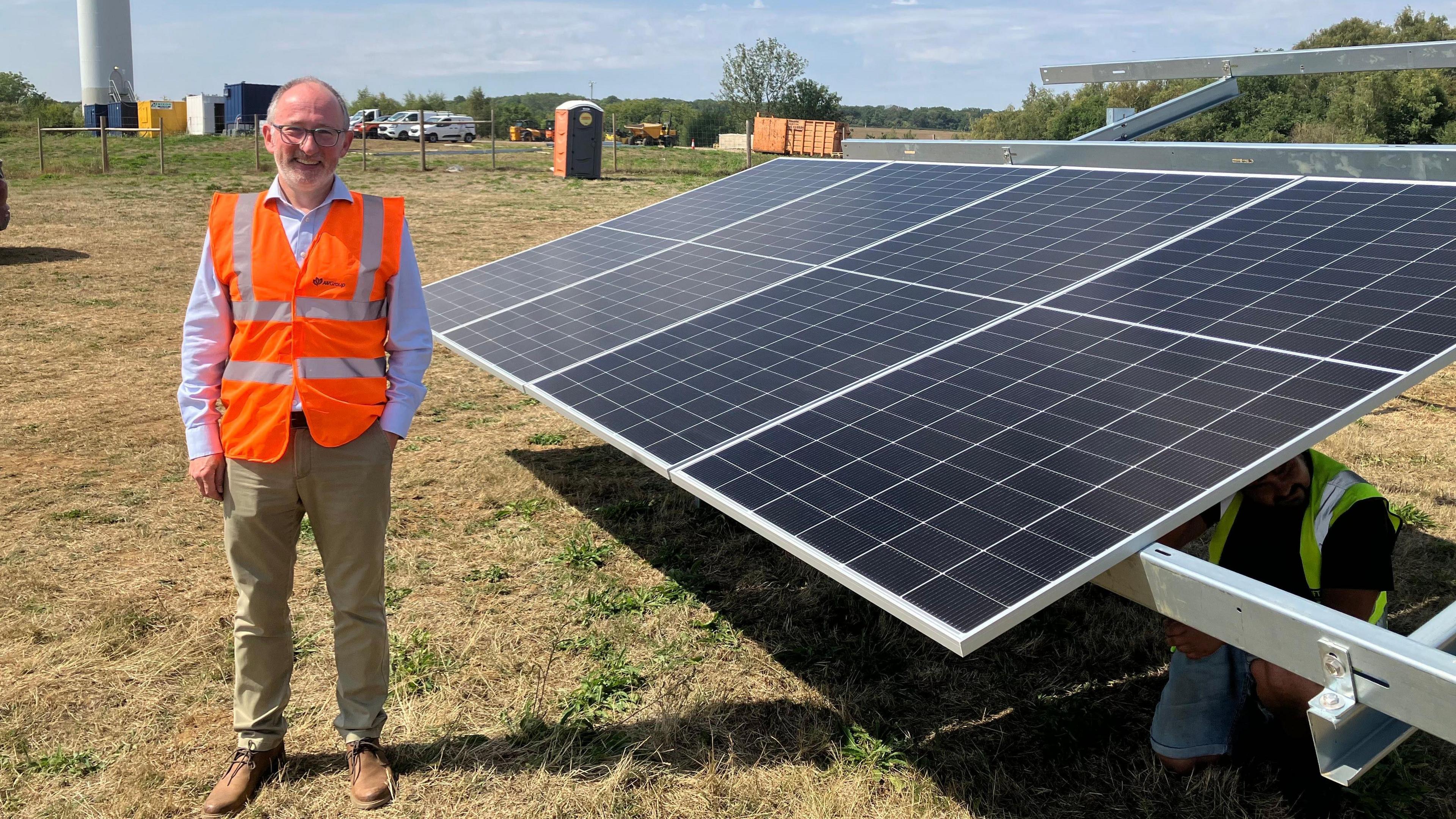 A picture of a man stood next to a solar panel, with a wind turbine behind him. He is wearing an orange high visibility jacket and has light green trousers on.