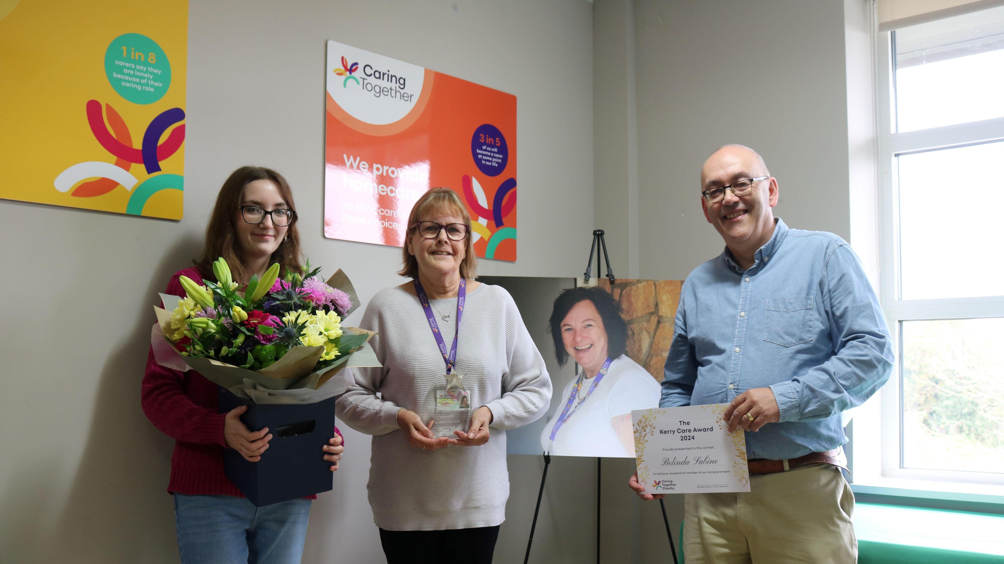 On the left is Megan Giles-Brown, daughter of the late Kerry Giles-Brown, holding a large bouquet of flowers. She has long brown hair, a purple jumper and blue jeans. She is wearing spectacles. Next to her, in the centre, is Belinda Sabine, with short sandy hair and glasses. She has a grey jumper on with a purple ribbon lanyard around her neck, and she is holding a small glass award. On the right is Steve Giles-Brown, Kerry's husband. He is bald with glasses and is wearing a blue shirt with beige trousers. He is holding a certificate